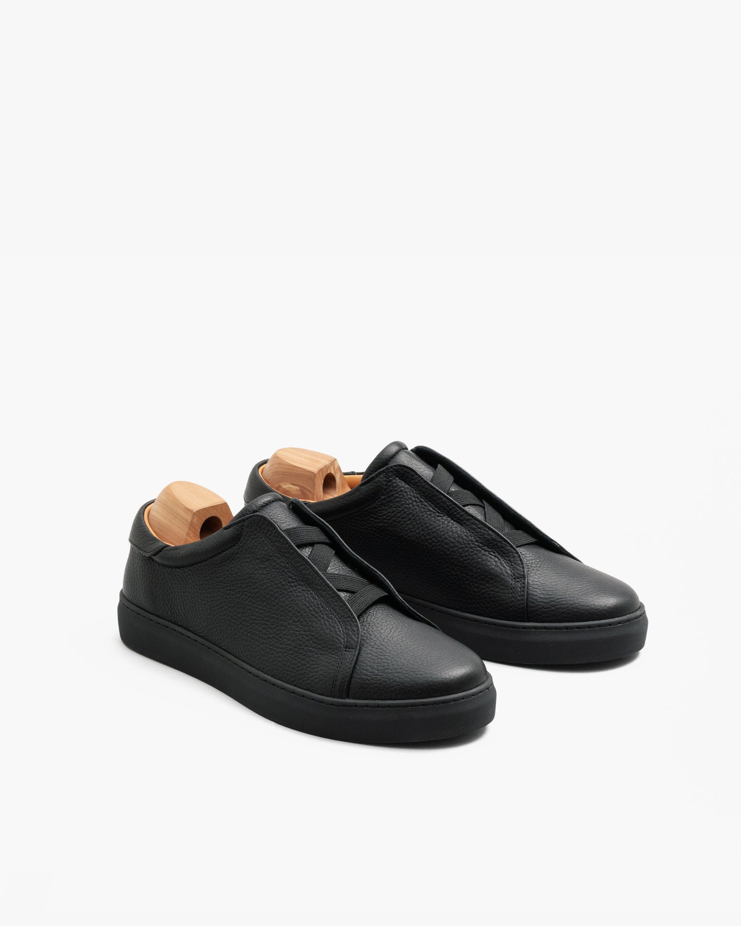 Alnö Monochrome II sneakers in black natural grain leather displayed at an angle, featuring a seamless monochrome look with matching black soles and laces, and wooden shoe trees inside each shoe.