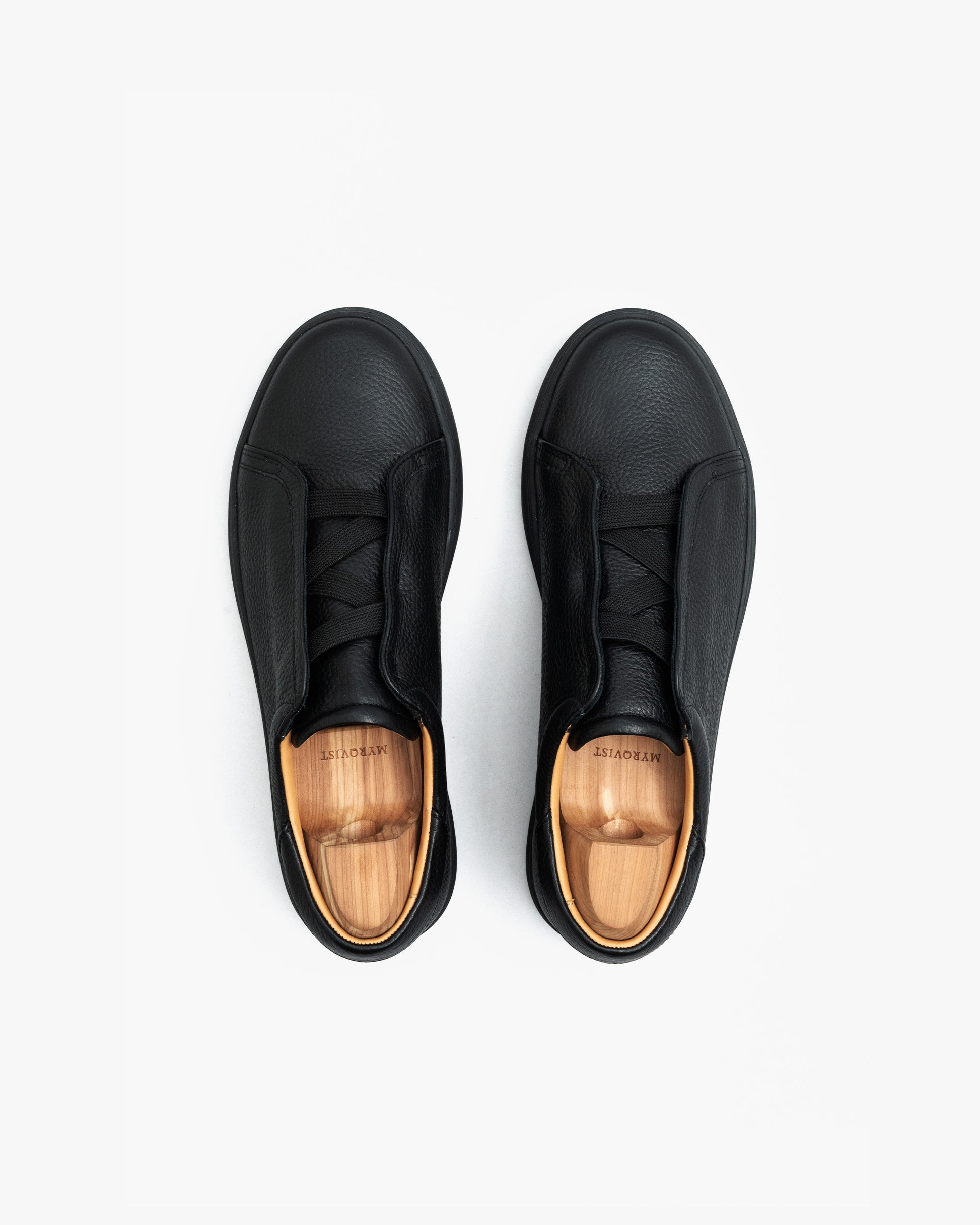 Pair of Alnö Monochrome II sneakers in black natural grain leather shown from above, highlighting the seamless monochrome design, textured finish, and wooden shoe trees inside each shoe.