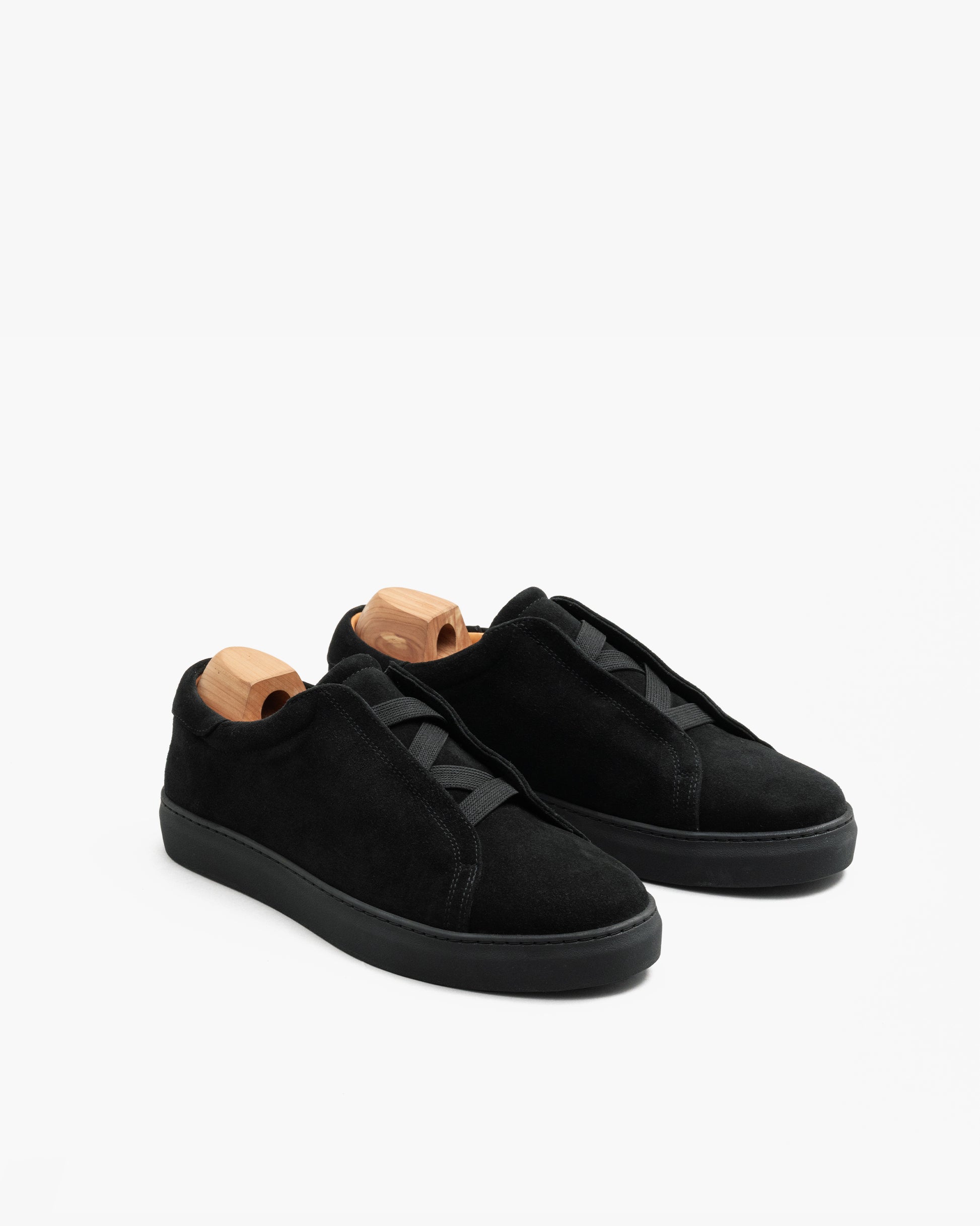 Alnö Monochrome II sneakers in black suede displayed at an angle, featuring seamless uppers, elastic lacing, and monochrome rubber soles with wooden shoe trees inside each shoe.