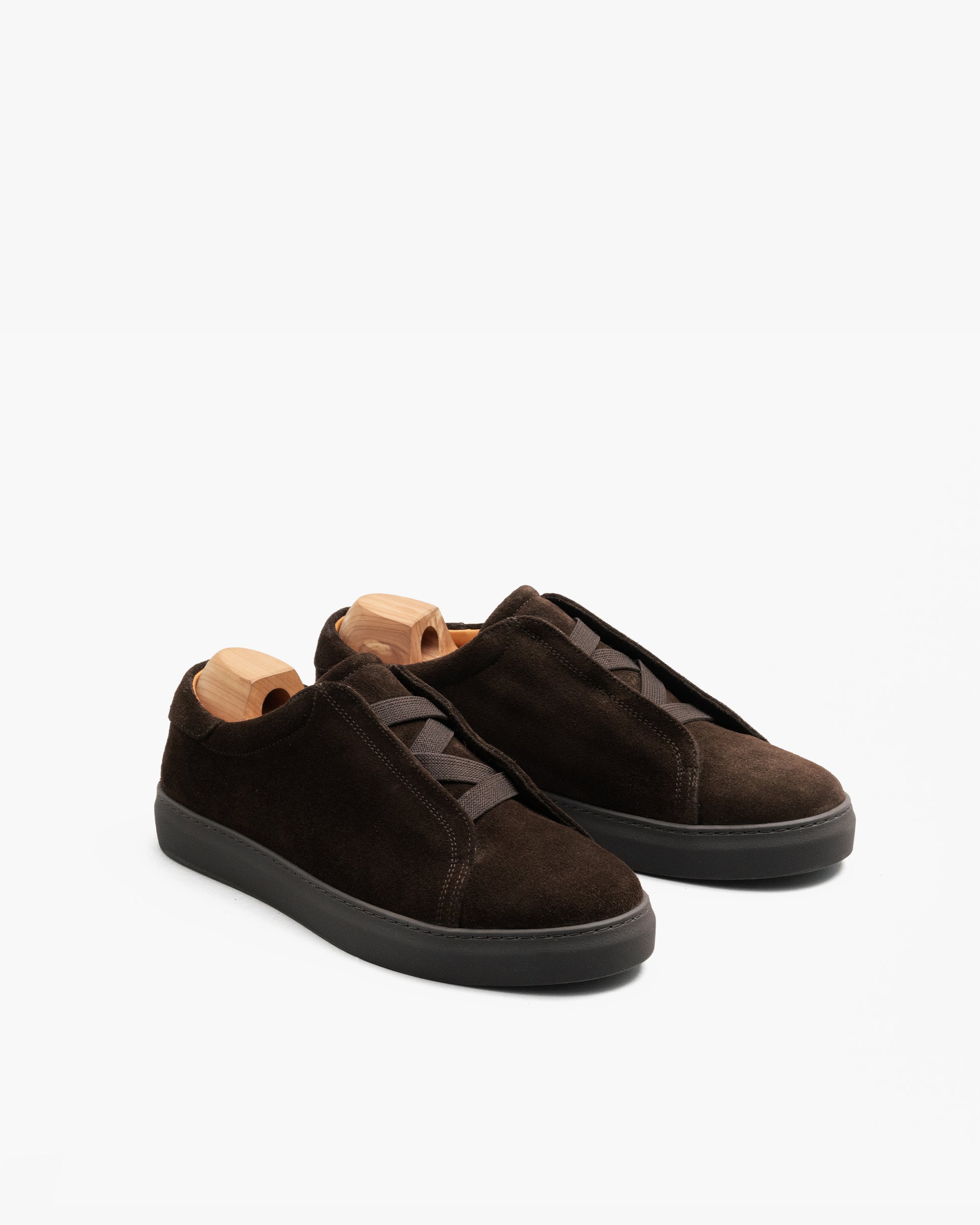 Alnö Monochrome II slip-on sneakers in dark brown suede with a monochrome rubber sole, tonal stitching, and elastic instep bands, displayed at an angle with wooden shoe trees inside.