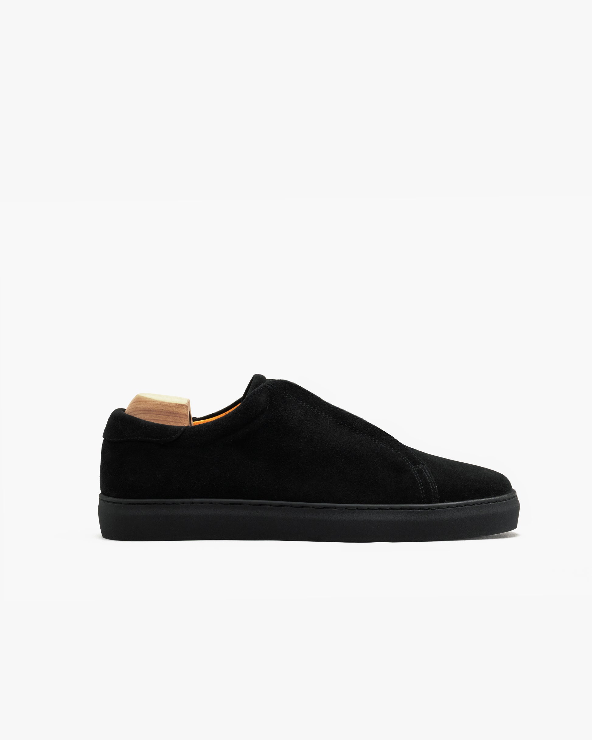 Low-top Alnö Monochrome II sneaker in black suede with a seamless upper, tonal stitching, and a monochrome rubber sole, shown in profile with a wooden shoe tree inside.