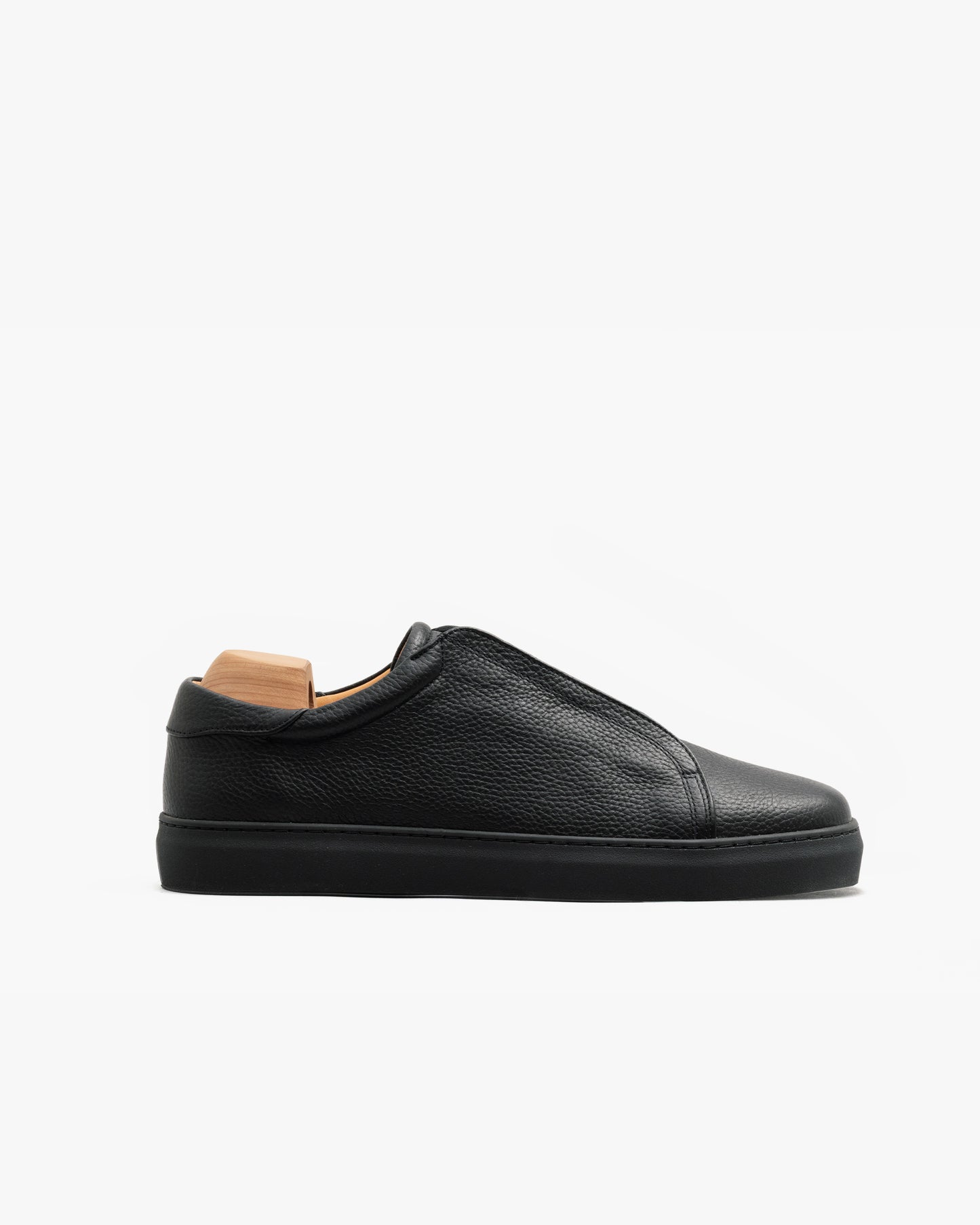 Minimalist black sneaker in textured natural grain leather with a tonal sole and laces, shown in profile on a white background with a wooden shoe tree inside.