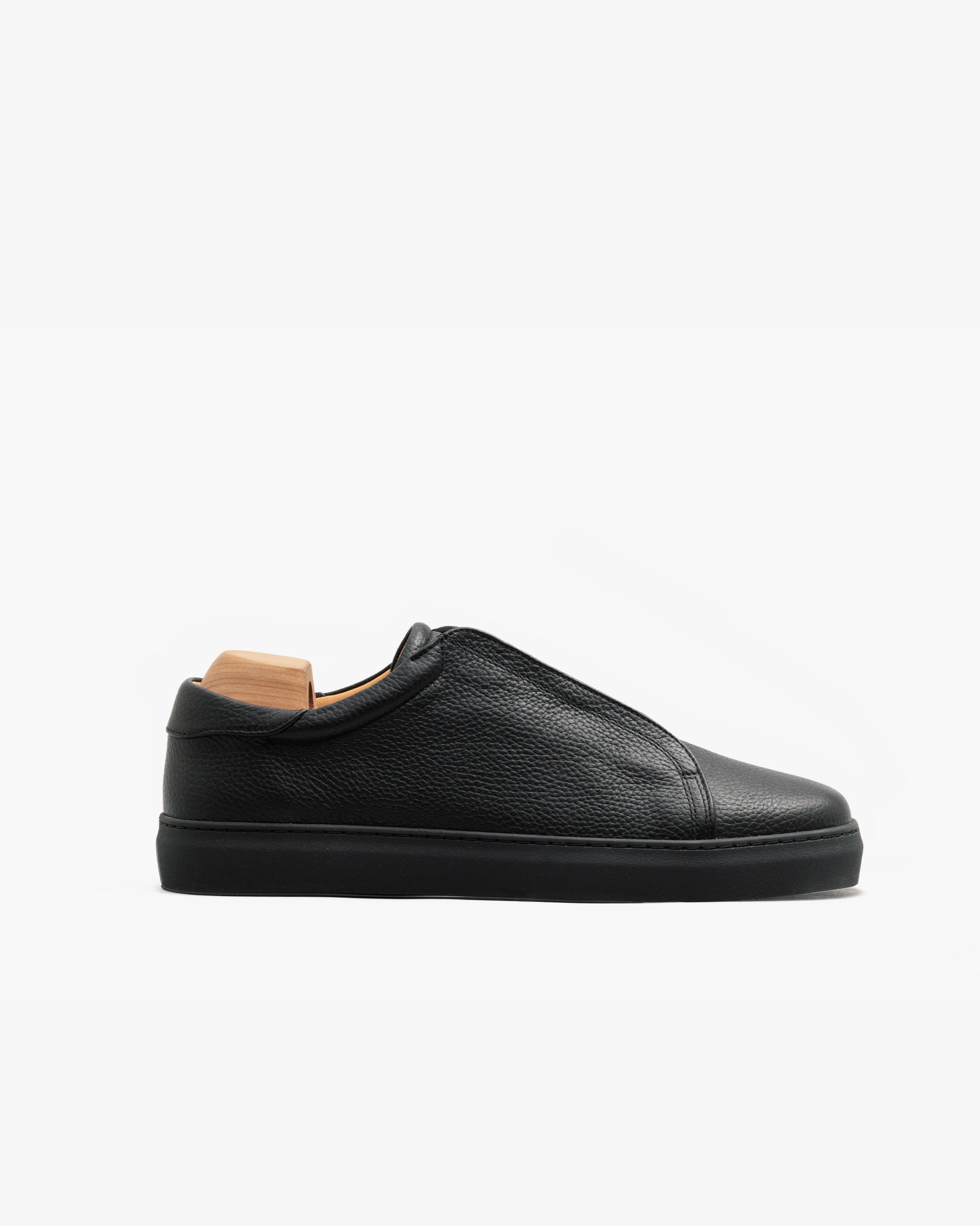 Minimalist black sneaker in textured natural grain leather with a tonal sole and laces, shown in profile on a white background with a wooden shoe tree inside.