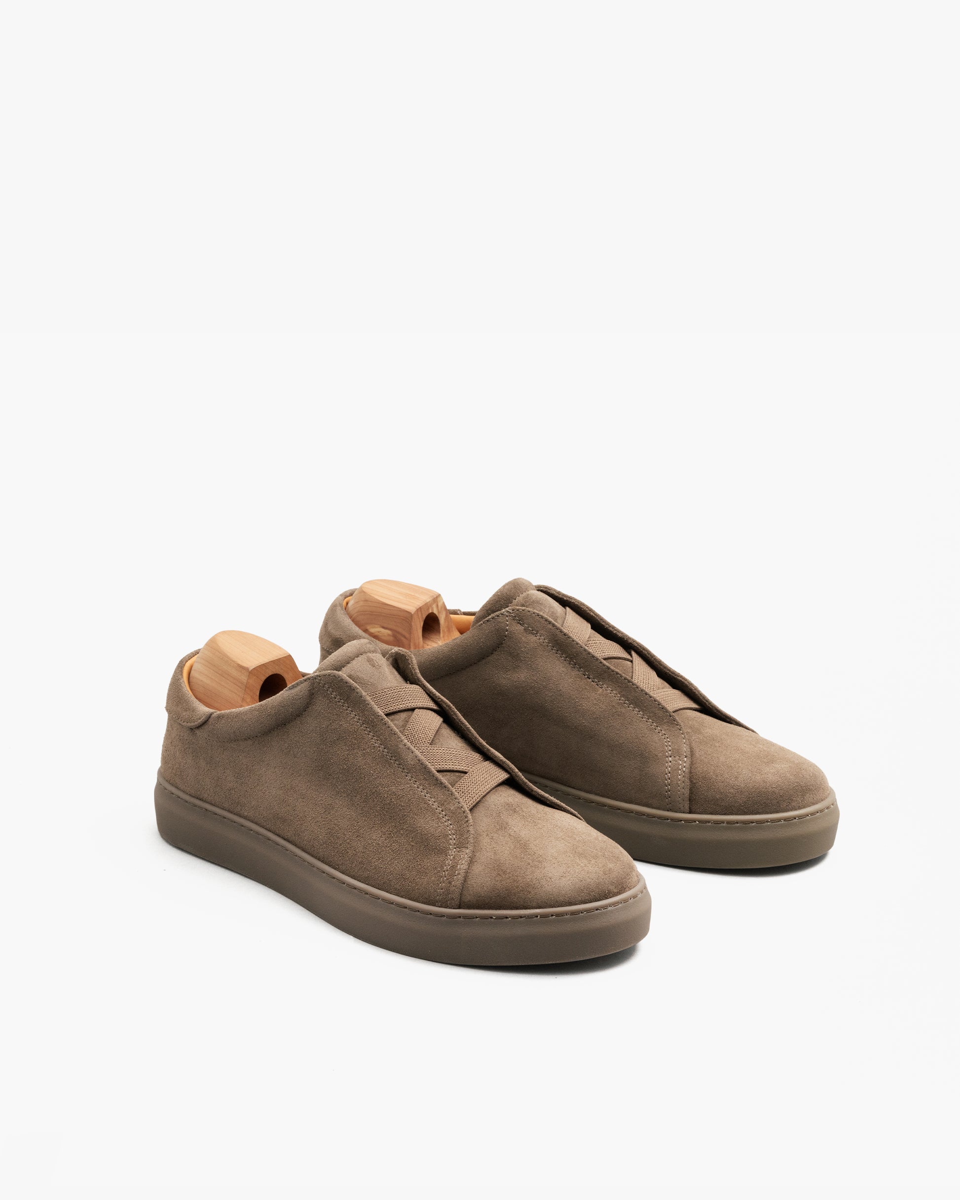 Alnö Monochrome II in taupe suede displayed at an angle, showing the soft suede upper, elastic side straps for easy slip-on wear, and color-matched rubber soles, with wooden shoe trees maintaining the shape.