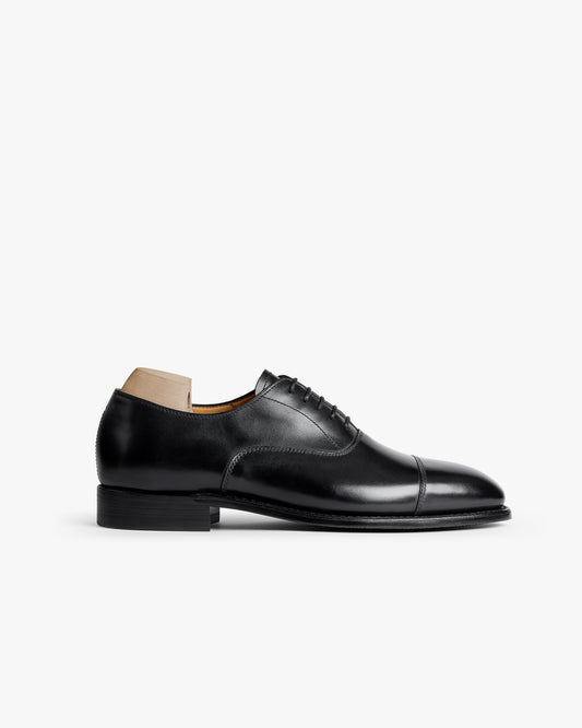 A cap-toe Oxford in black calf leather with a chiseled toe, shown in profile with a shoe tree against a white background.