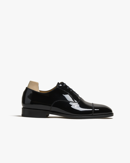 Black patent leather oxford shoe with a cap toe, five-eyelet lace system, and sleek silhouette, featuring a high-gloss finish and subtle stitching, ideal for formal occasions or pairing with a tuxedo.