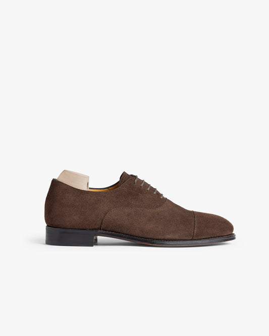 A side profile of Äppelviken II – Dark Brown Suede, a cap-toe oxford shoe with shoe tree inside, against a white background.