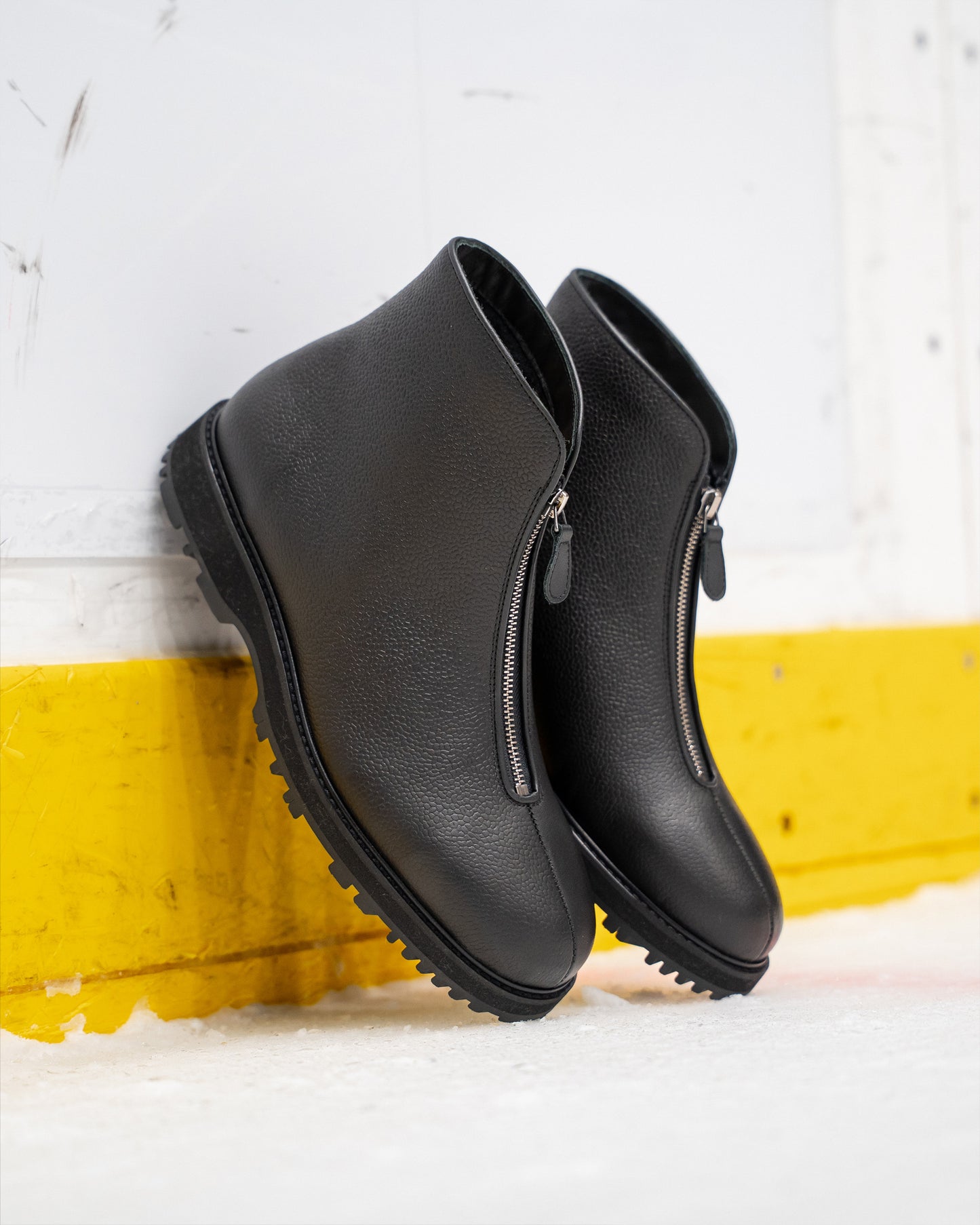 Pair of Funäs in black country calf boots with central zippers and textured full-grain leather, shown leaning against an ice rink barrier, highlighting their rugged Explorer soles and winter-ready design.