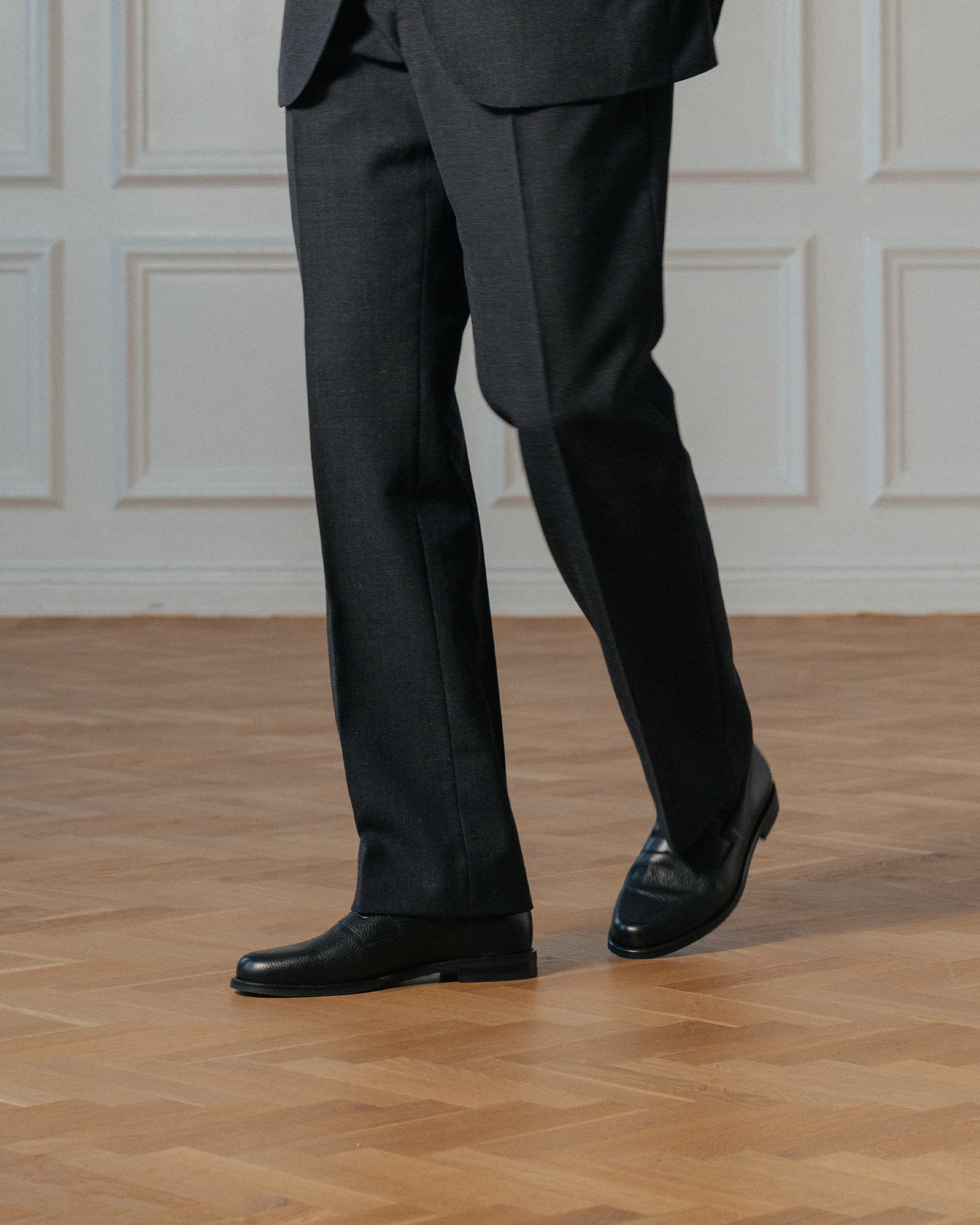 Black grain leather shoes worn with charcoal suit trousers on a herringbone parquet floor