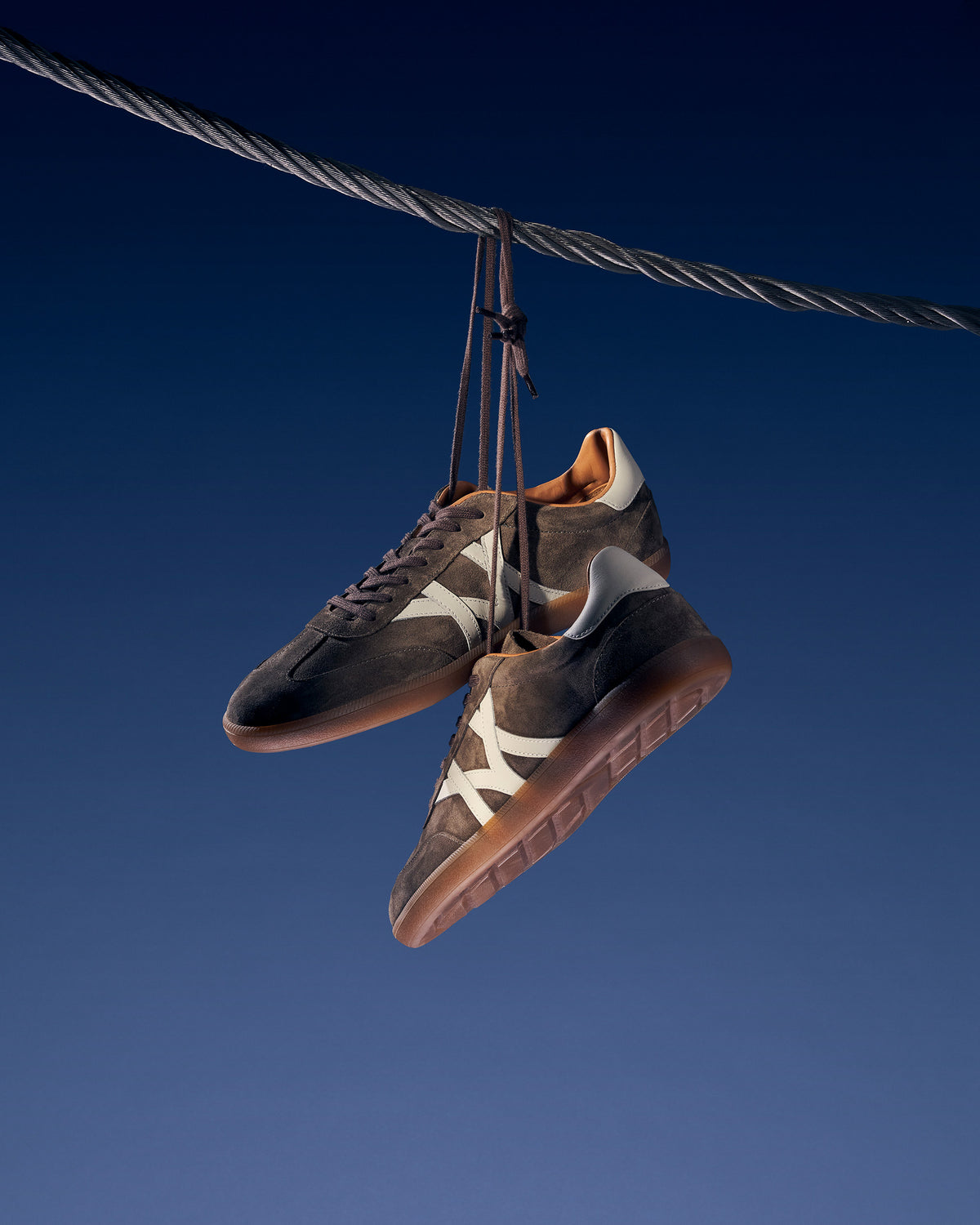 Högalid in soft taupe suede hanging by its laces from a wire against a deep blue background.