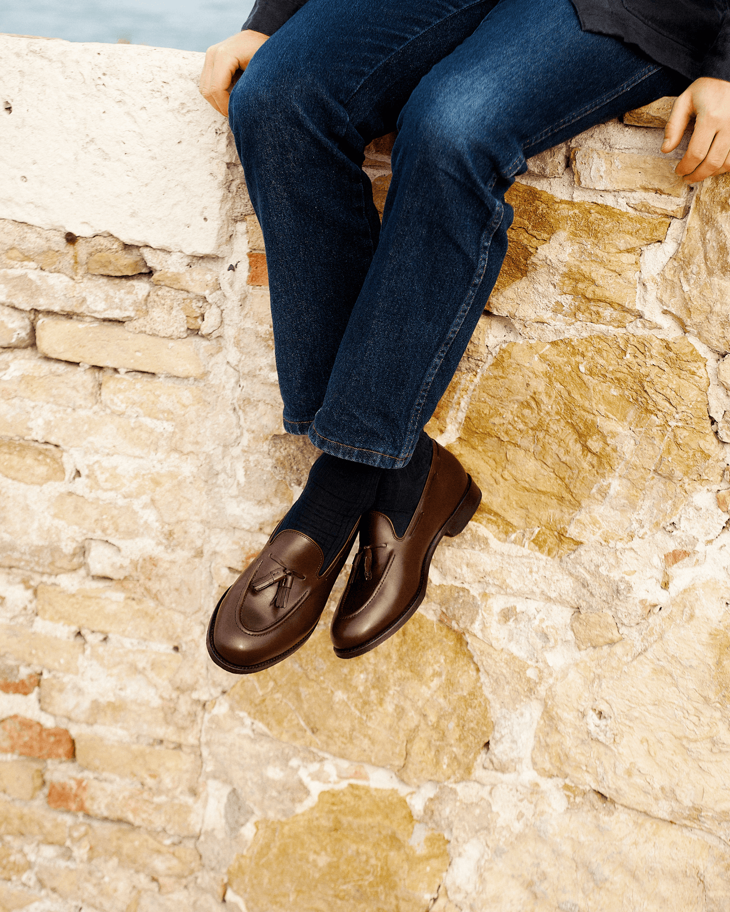 A person wearing Mölle II in dark brown calf sits on a stone wall, pairing the polished tassel loafers with dark socks and blue denim for a relaxed yet refined look.