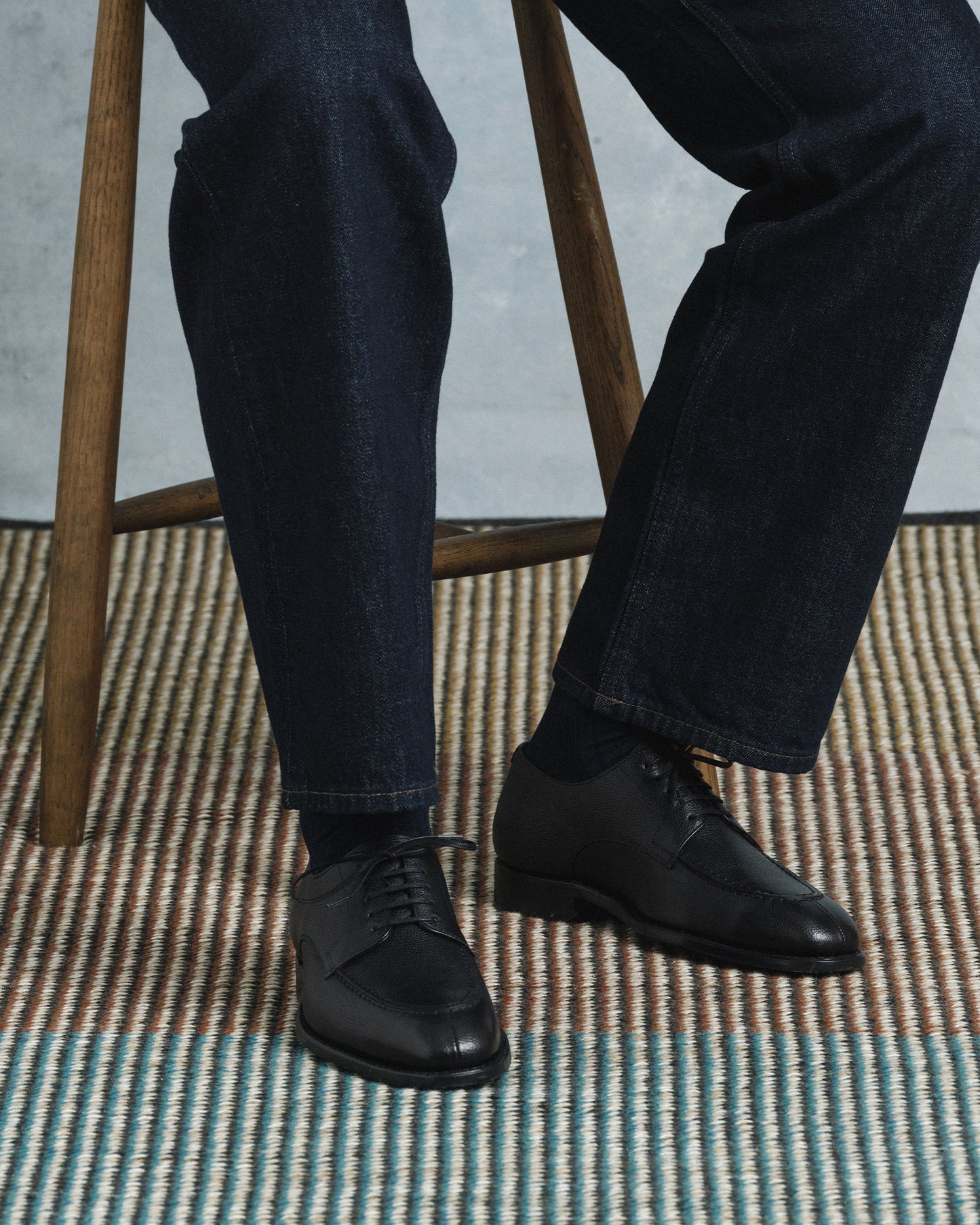 A man sits on a wooden stool wearing Norrmalm II in black hatch grain, styled with dark denim trousers, showcasing the textured split toe derby shoes in a relaxed, modern setting.