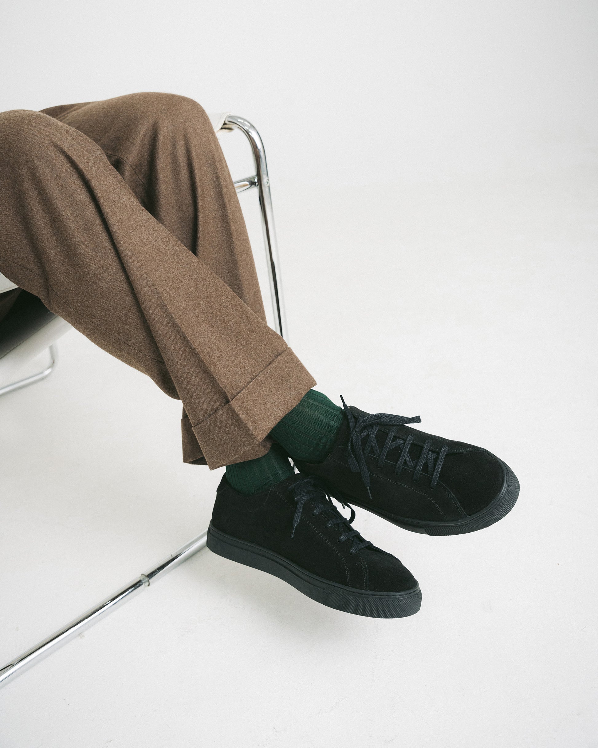 A person sitting down wearing Oaxen Monochrome II – Black Suede sneakers with brown trousers and green socks.