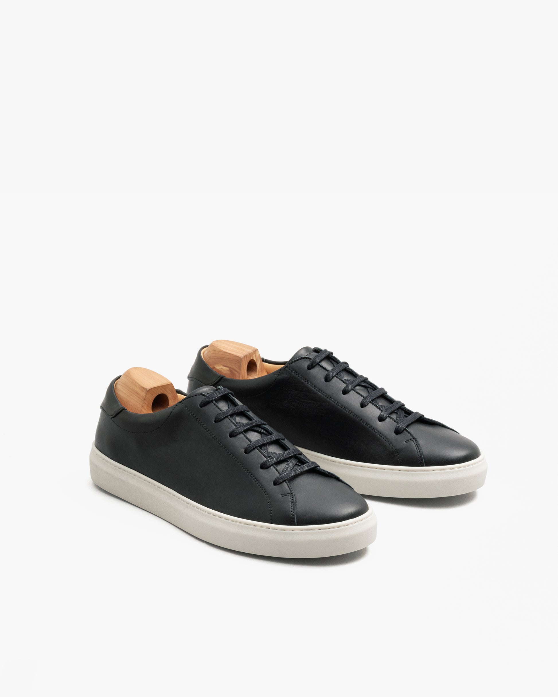 Oaxen in black calf leather shown at an angle, highlighting the smooth black upper, crisp black laces, white M-pattern sole, and wooden shoe trees for shape retention.