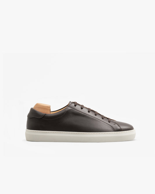 Oaxen in dark brown calf features a low-profile silhouette with supple brown leather, precise stitching, and a crisp white rubber sole, shown with a wooden shoe tree against a clean white background.