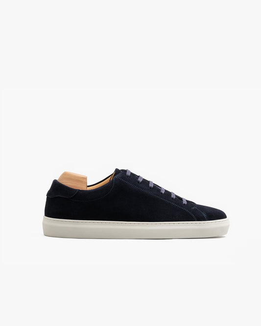 Low-top men’s sneaker in navy suede with tonal stitching, flat cotton laces, and a softly rounded toe, set on a crisp off-white sole and displayed with a wooden shoe tree.
