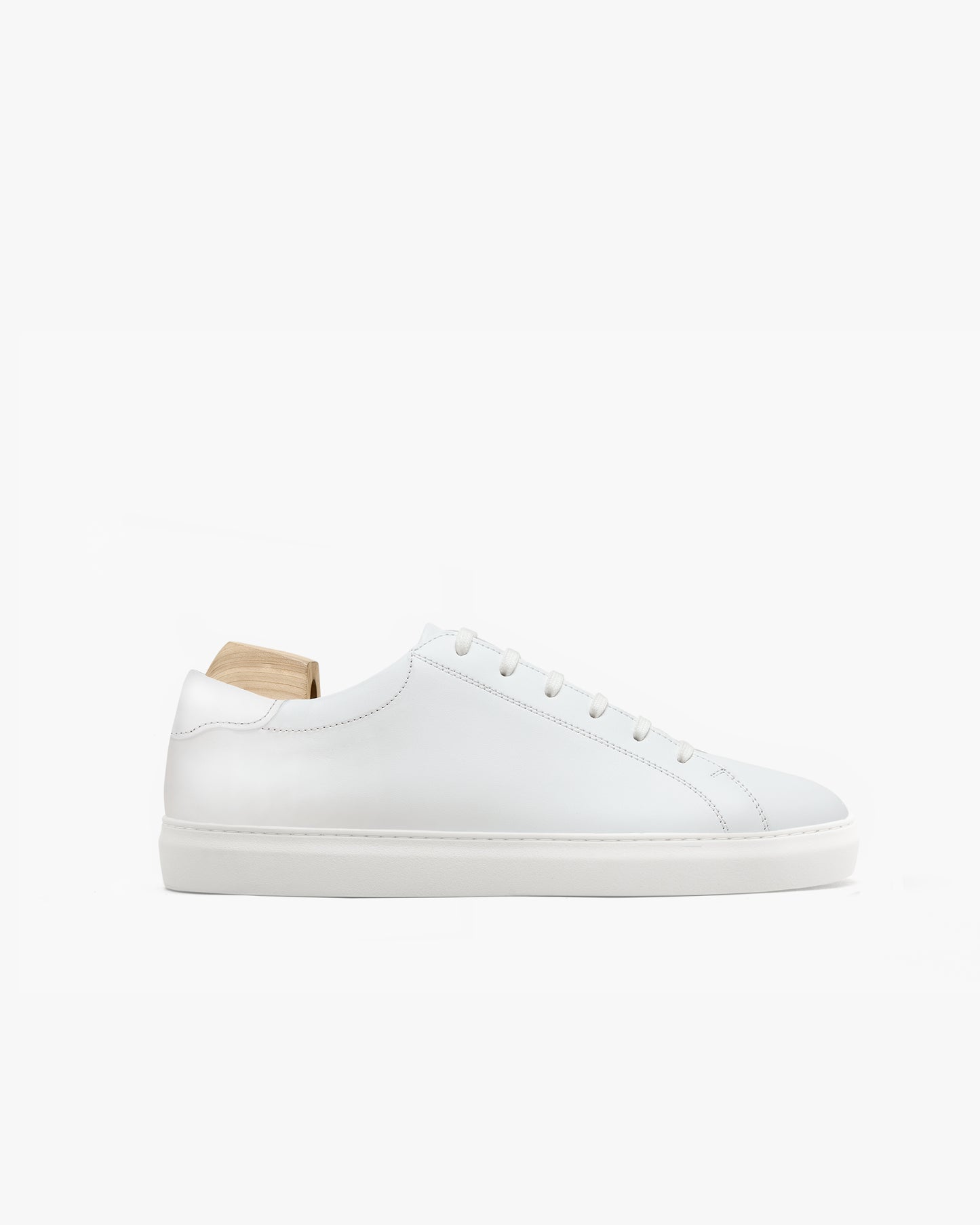 Minimalist white leather sneaker with tonal laces and subtle stitching, shown in profile with a wooden shoe tree inside, highlighting its clean lines and softly rounded toe.