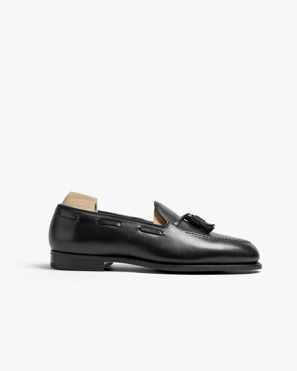 Mölle II in black calf displayed in profile, showcasing its smooth leather upper, apron front, and tassel detailing, with a single leather sole and wooden shoe tree inserted.