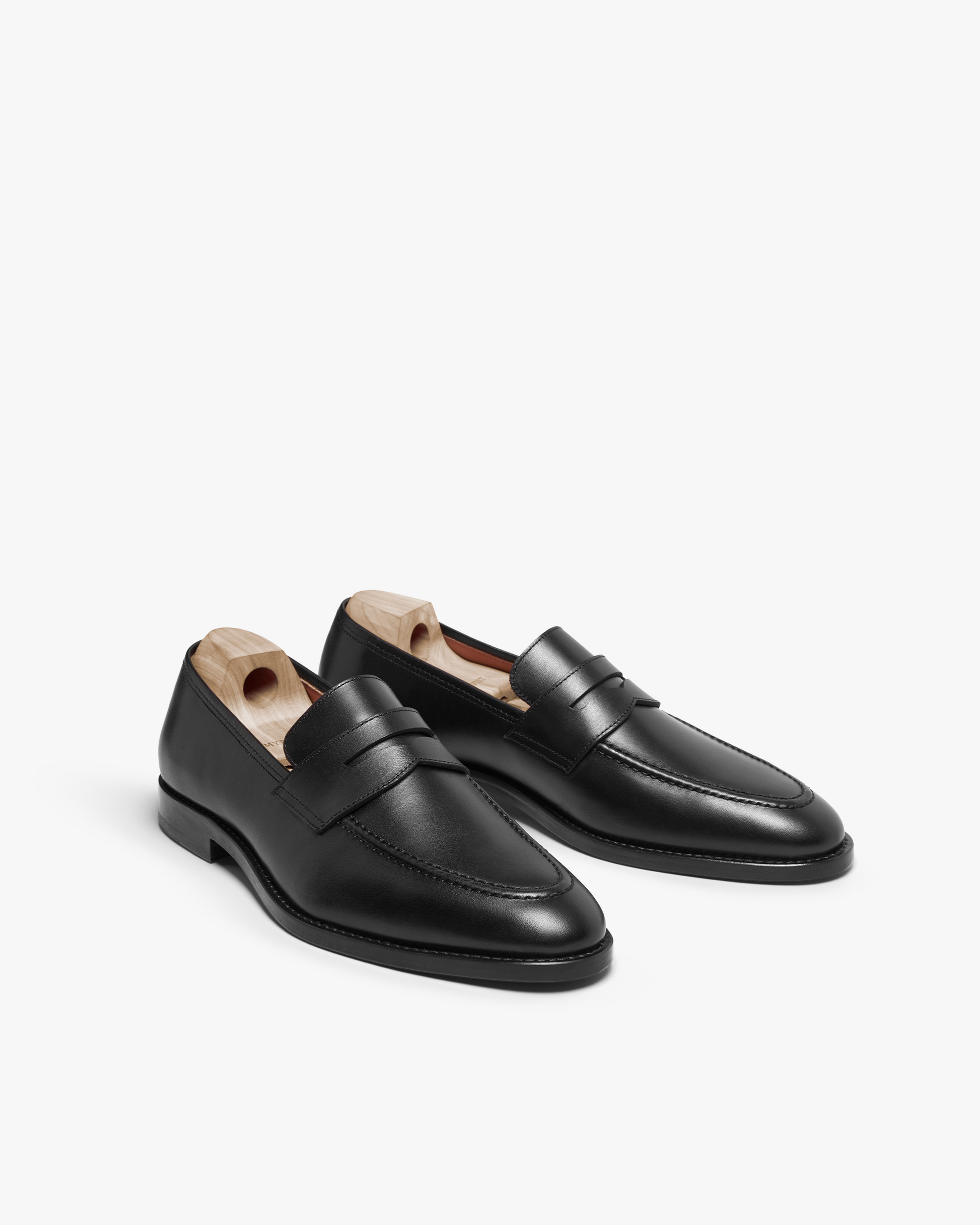Smögen loafers in black calf leather, presented at an angle to showcase the sleek penny strap design, gently rounded toe, and subtle sheen, each fitted with a wooden shoe tree.