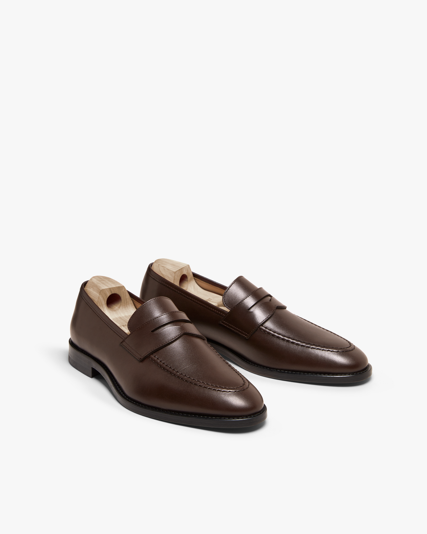 Smögen penny loafers in dark brown calf leather, featuring smooth contours, refined stitching, and gently rounded toes, displayed with wooden shoe trees for shape retention.