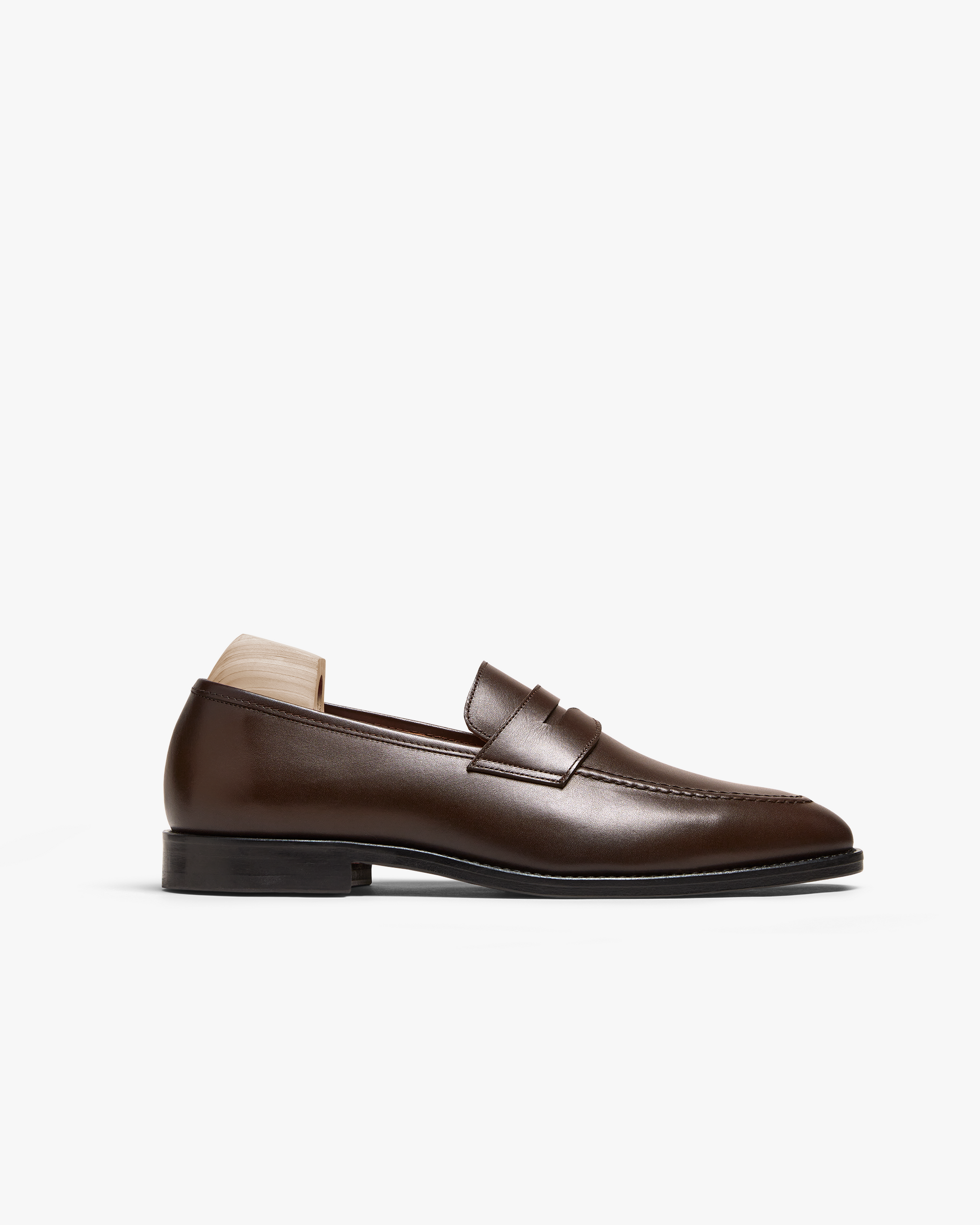 Dark brown calf penny loafer with smooth full-grain leather, gently rounded toe, subtle stitching, and a black sole, shown in profile with a wooden shoe tree inserted.