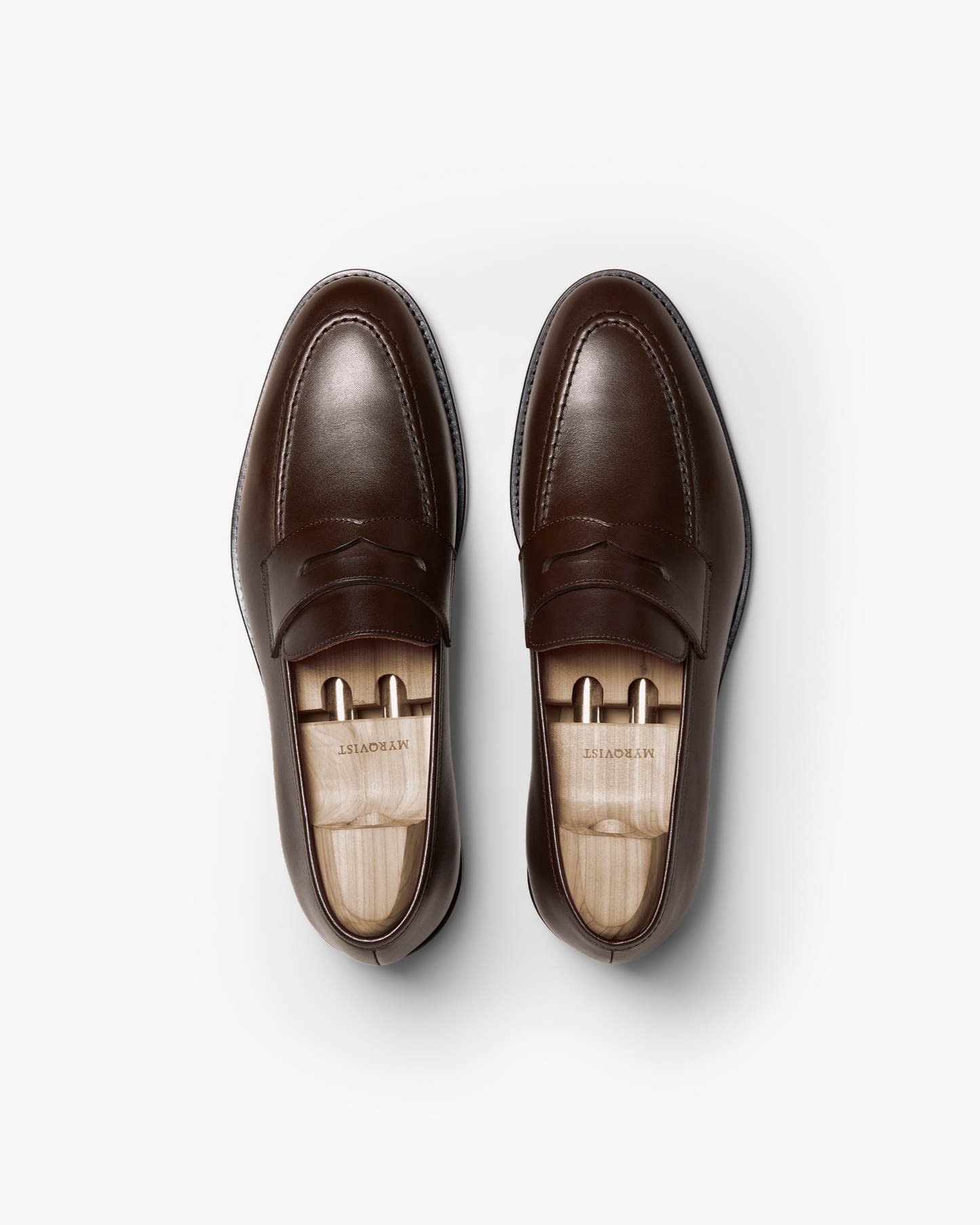 Pair of Smögen penny loafers in dark brown calf leather, shown from above with wooden shoe trees, highlighting the smooth finish, gently rounded toe, and refined stitching.