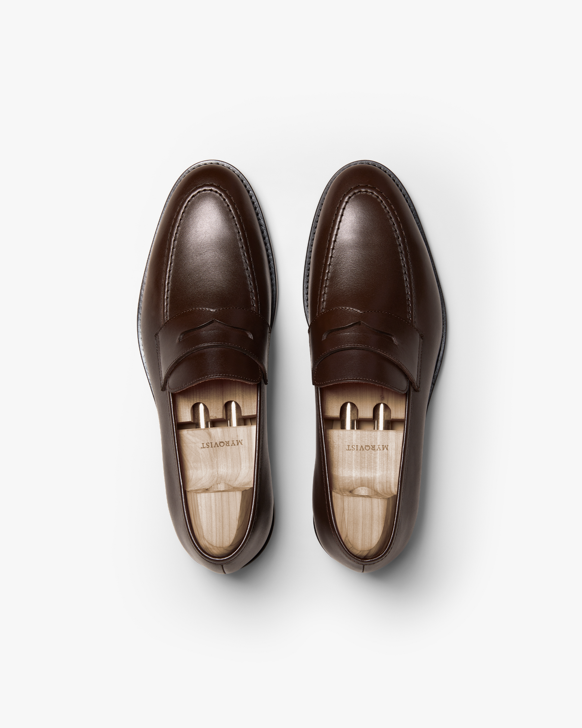 Pair of Smögen penny loafers in dark brown calf leather, shown from above with wooden shoe trees, highlighting the smooth finish, gently rounded toe, and refined stitching.