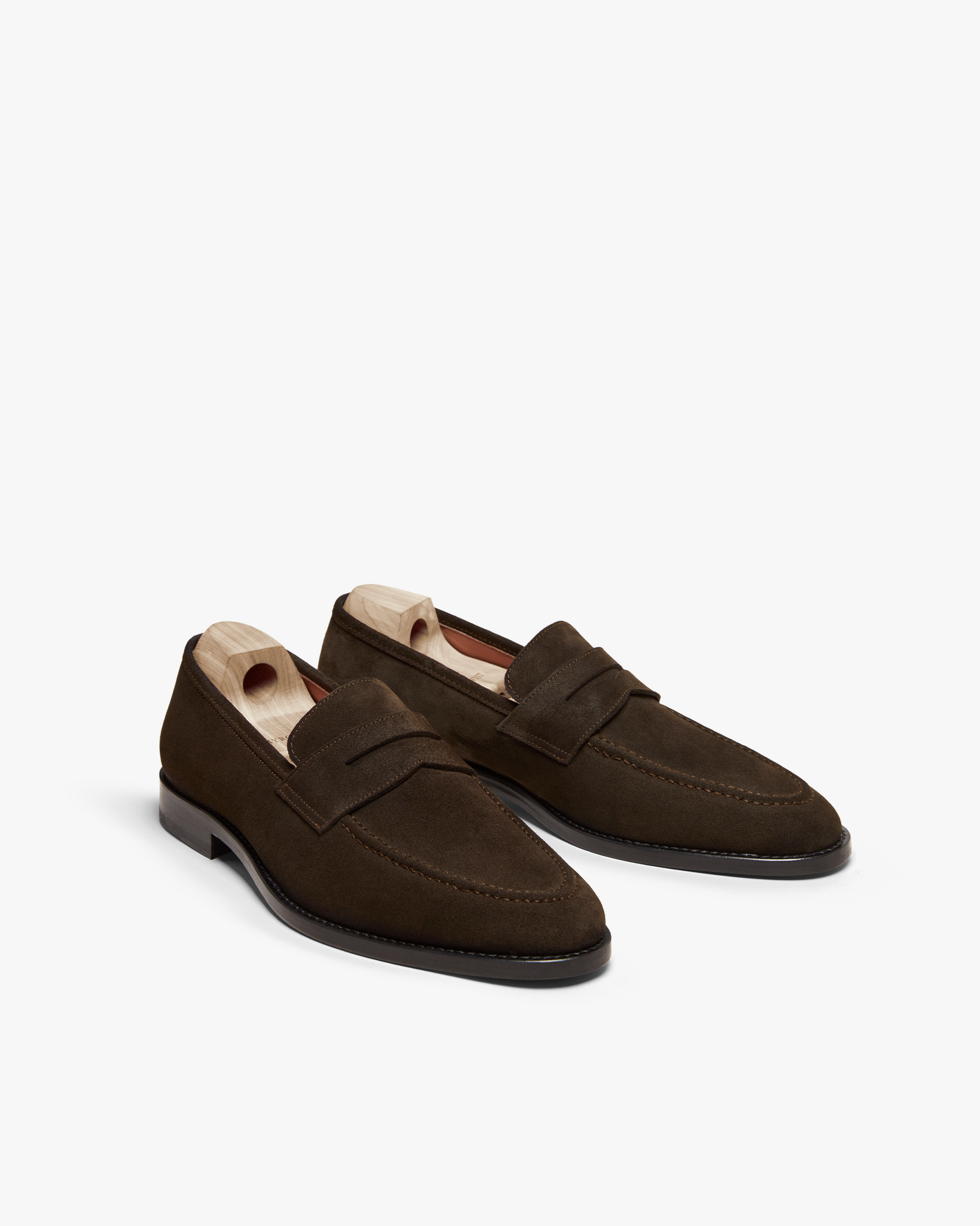 Smögen loafers in dark brown suede, featuring a classic penny strap, gently rounded toe, and fine stitching, presented with wooden shoe trees to maintain their elegant shape.