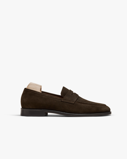 Dark brown suede penny loafer with a gently rounded toe, subtle stitching, and a black sole, shown in profile with a wooden shoe tree supporting the shape.