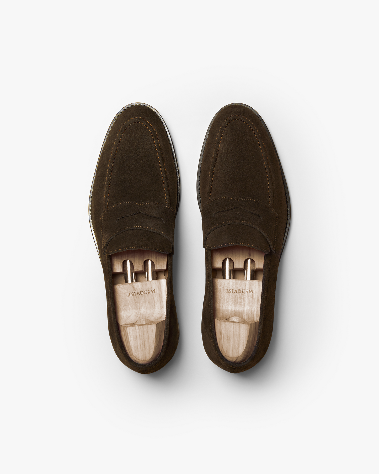 Pair of Smögen loafers in dark brown suede, shown from above with wooden shoe trees, highlighting the penny strap detail, fine stitching, and soft suede texture.