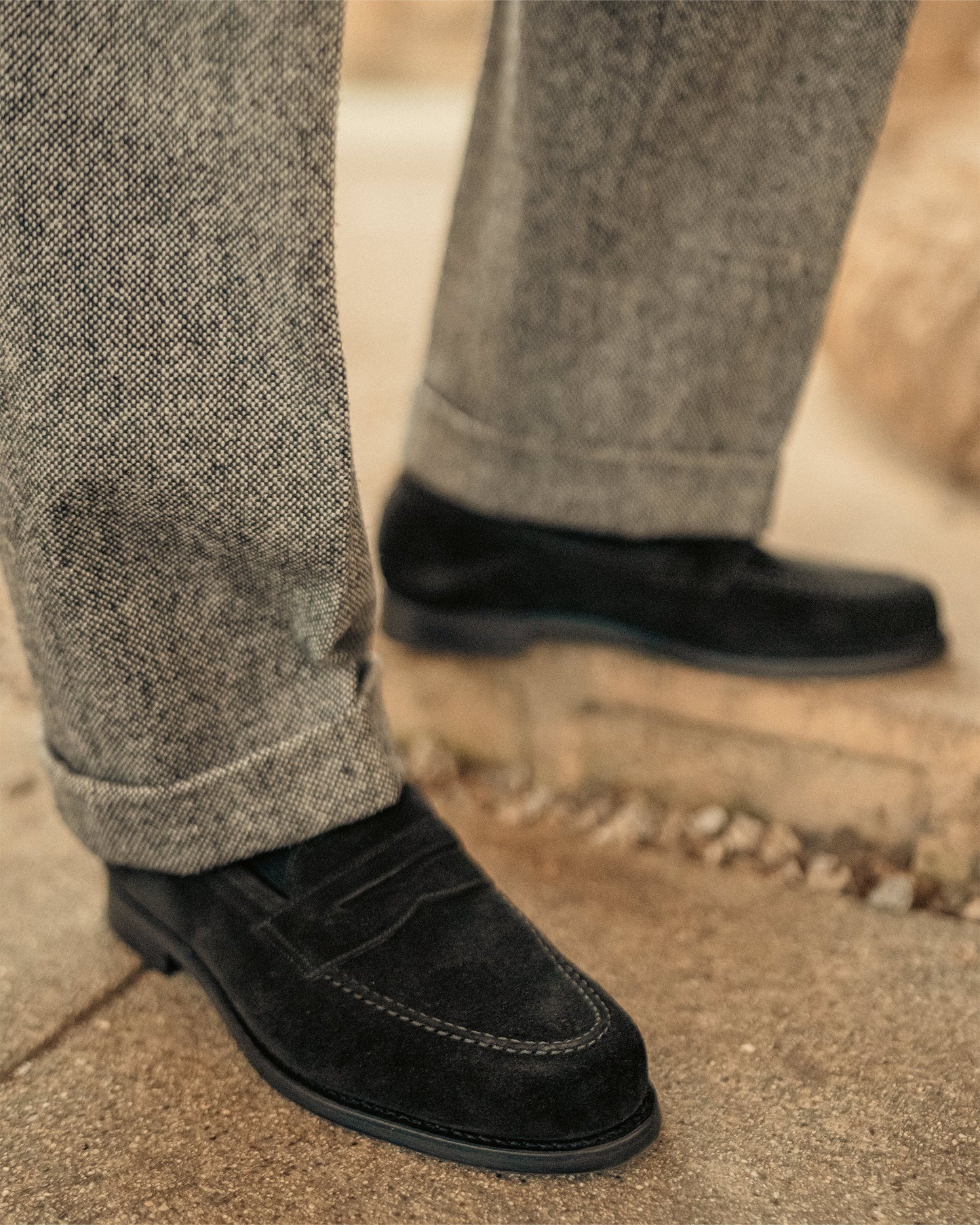 Close-up of Stenhammar II in black suede penny loafers worn with cuffed gray slacks, highlighting the soft suede texture, apron stitching, and versatile style for refined or relaxed settings.
