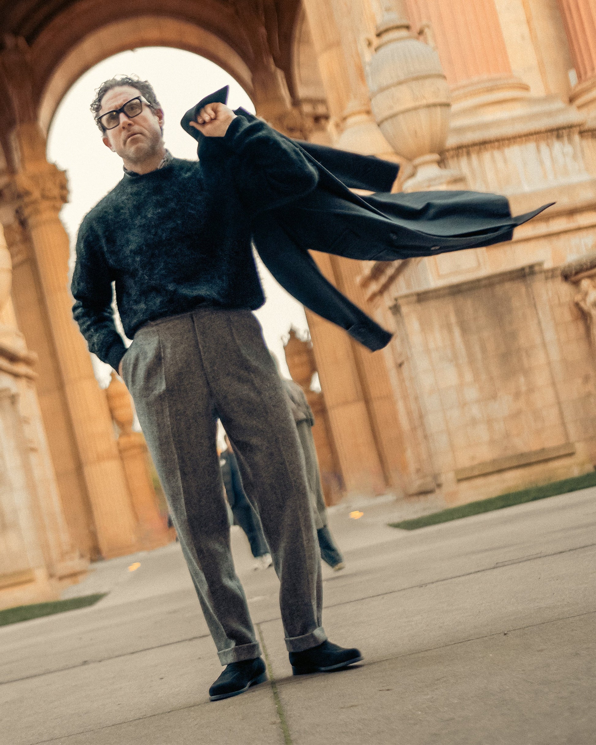A man wearing Stenhammar II in black suede penny loafers styled with tailored gray slacks and a dark sweater, standing outdoors in front of grand architectural columns for a refined, city-ready look.