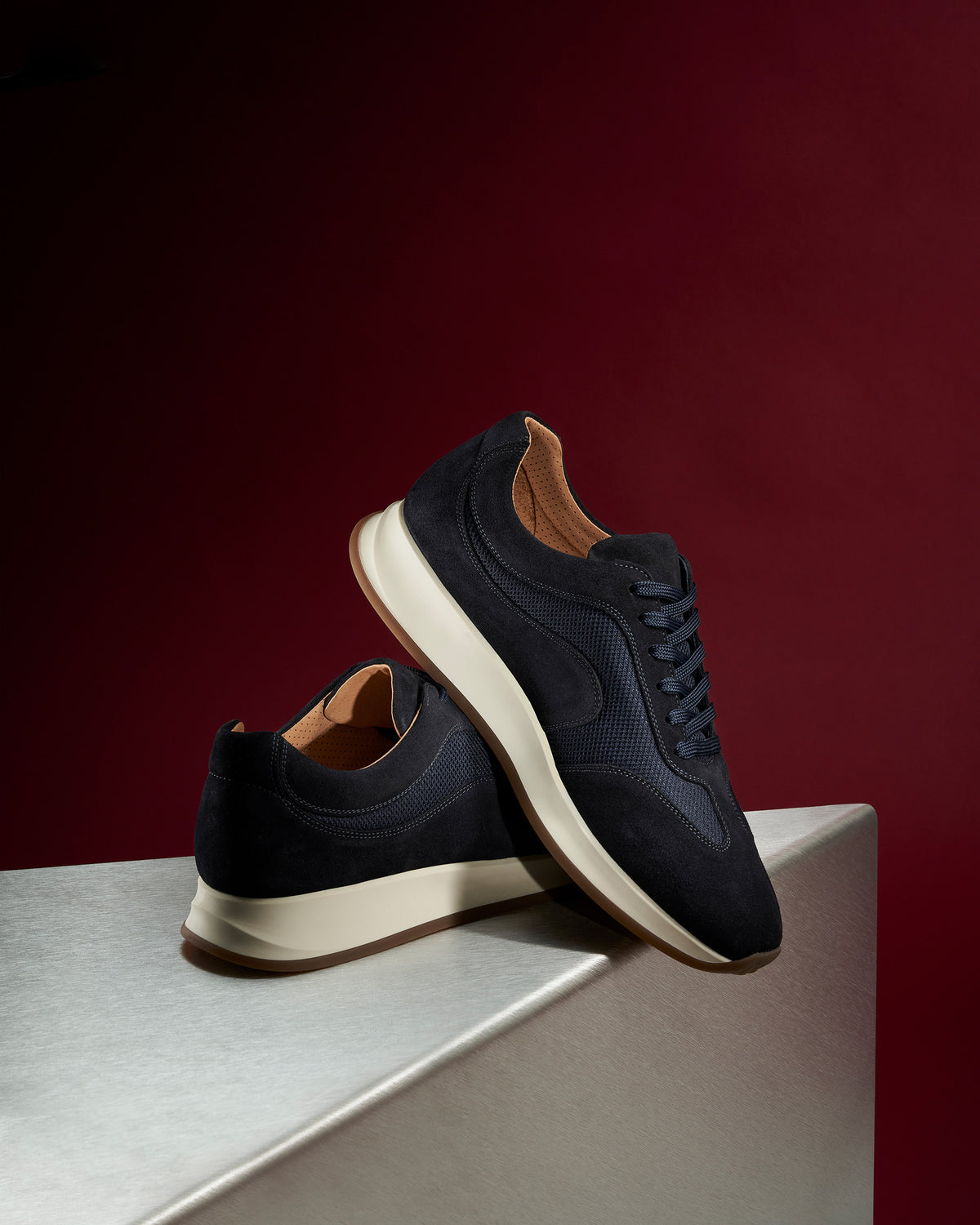 Stensund II – Navy Suede sneakers displayed on a metallic pedestal against a dark red background.