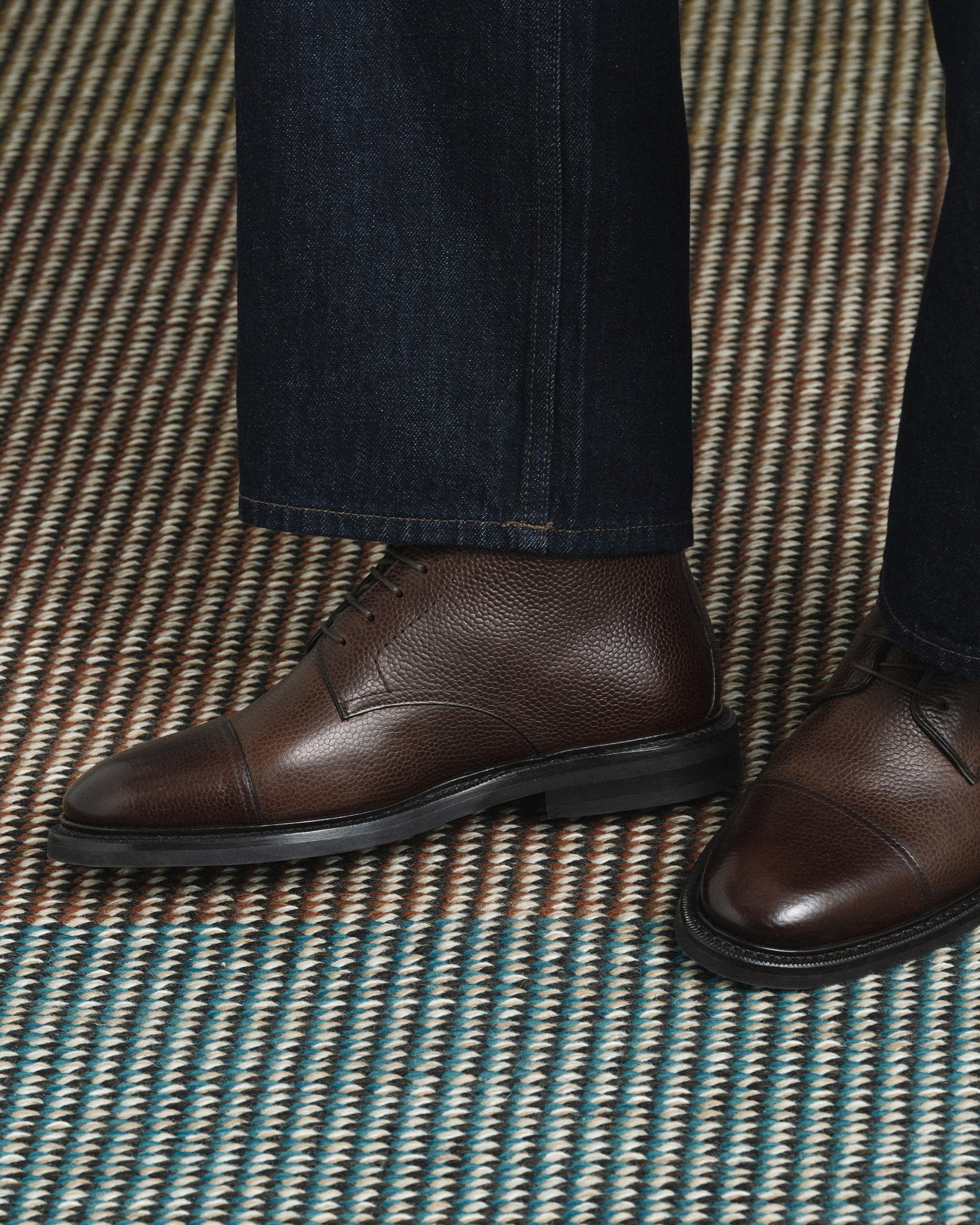Abisko in dark brown country calf is shown worn with dark denim, highlighting the textured grained leather, cap toe design, and sturdy rubber sole for a refined yet versatile look suitable for both casual and smart settings.