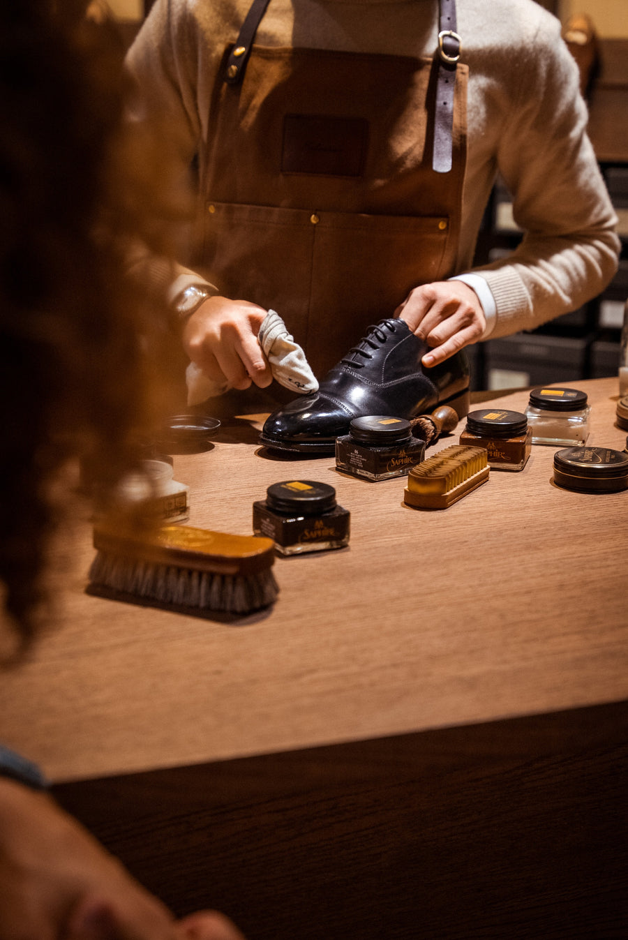 Hands in an apron work shoe cream into a black leather oxford with a cloth, with open tins of cream on the counter.