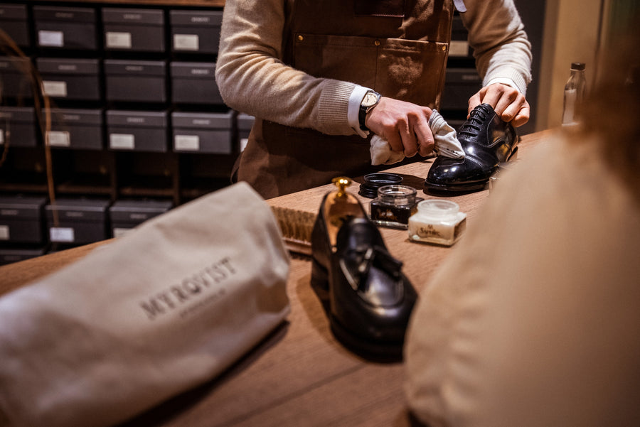 A person in an apron works a white cloth over a black leather oxford at a store counter, with tins of polish and a brush beside it.