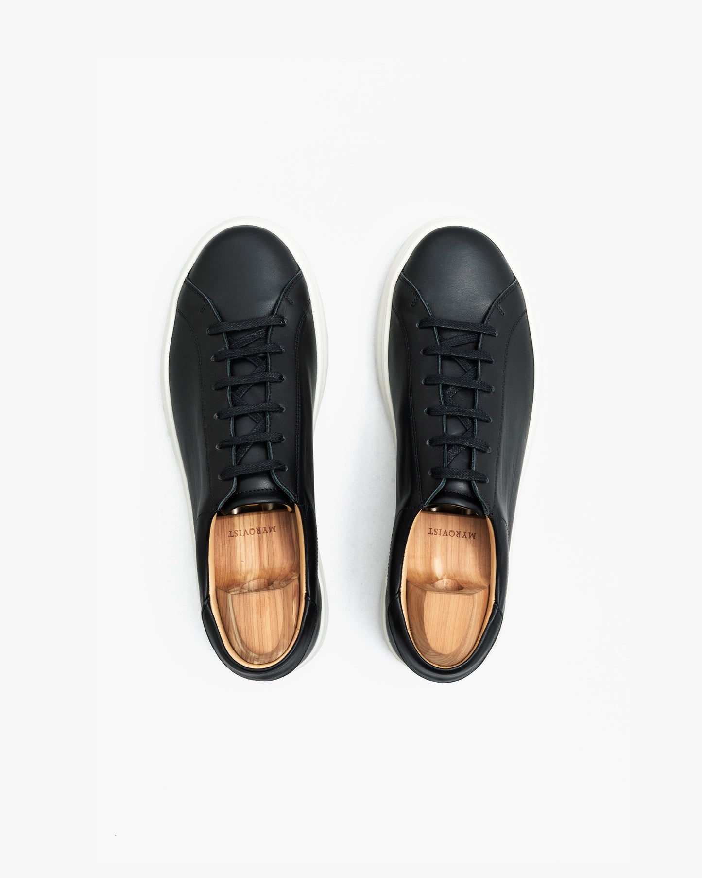 Pair of Oaxen sneakers in black calf leather shown from above, featuring clean stitching, black laces, white soles, and wooden shoe trees inside each shoe.