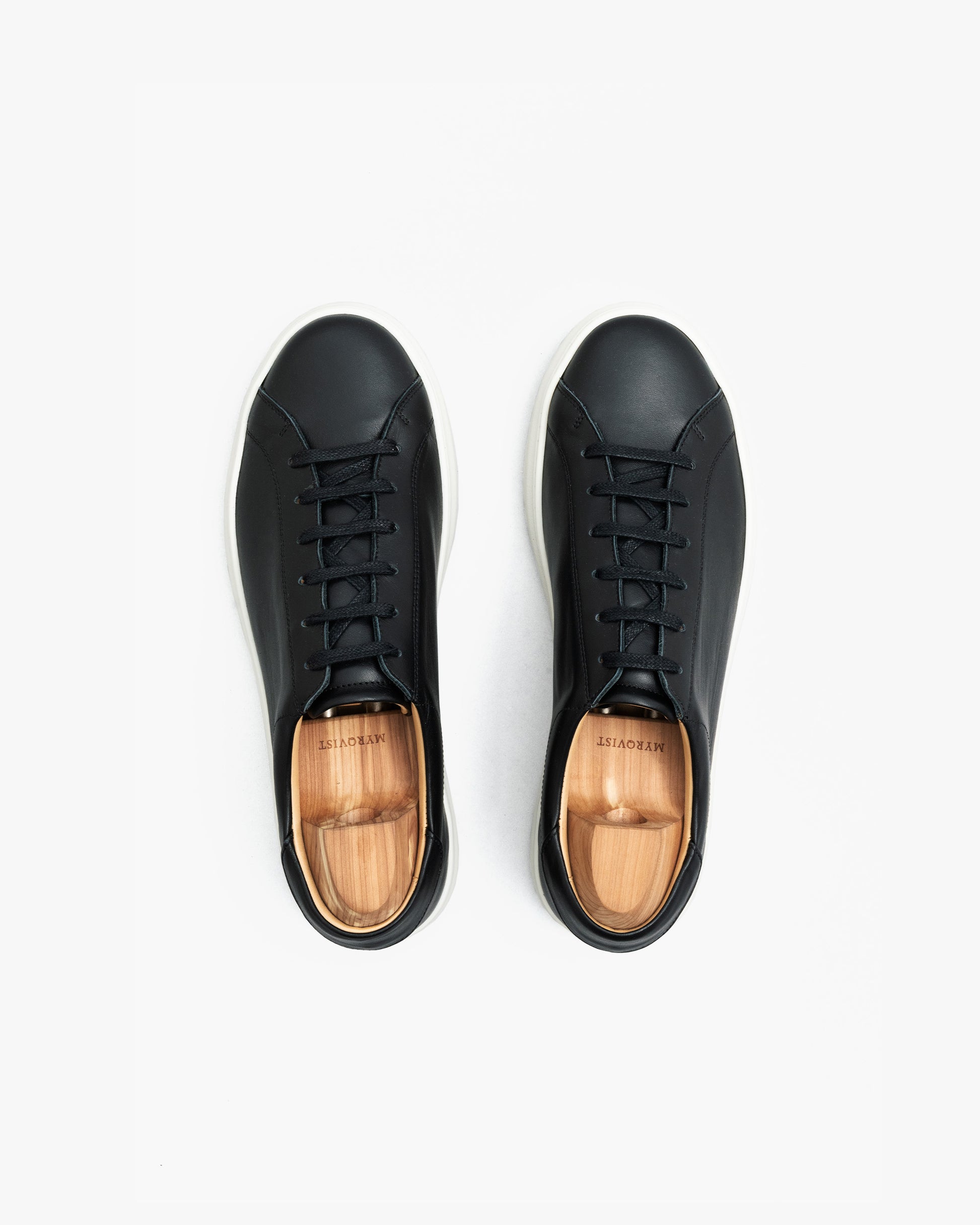 Pair of Oaxen sneakers in black calf leather shown from above, featuring clean stitching, black laces, white soles, and wooden shoe trees inside each shoe.