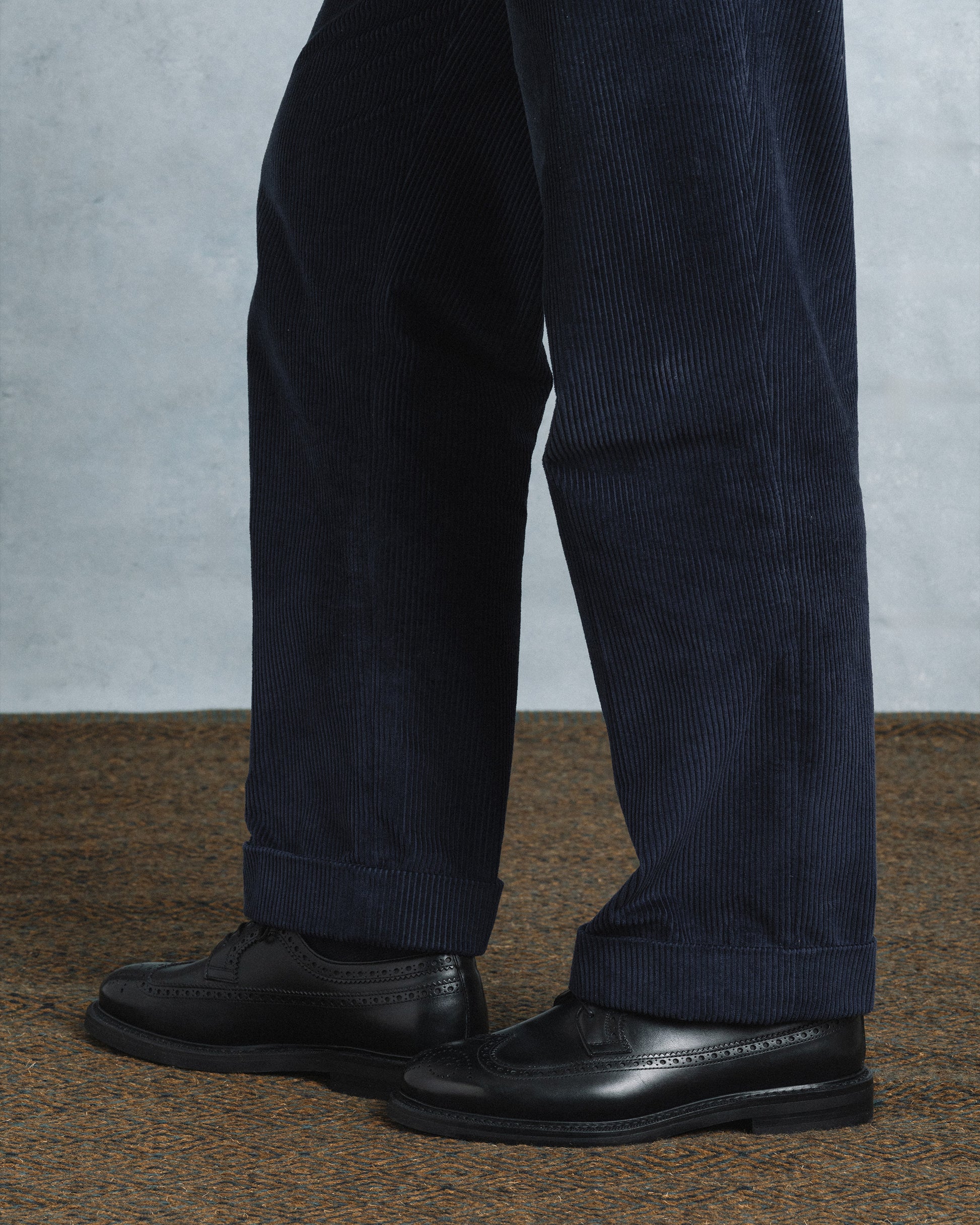 Orust II in black calf shown on foot with navy corduroy trousers, highlighting the brogue detailing, rounded toe, and sturdy studded sole for a refined yet versatile look.