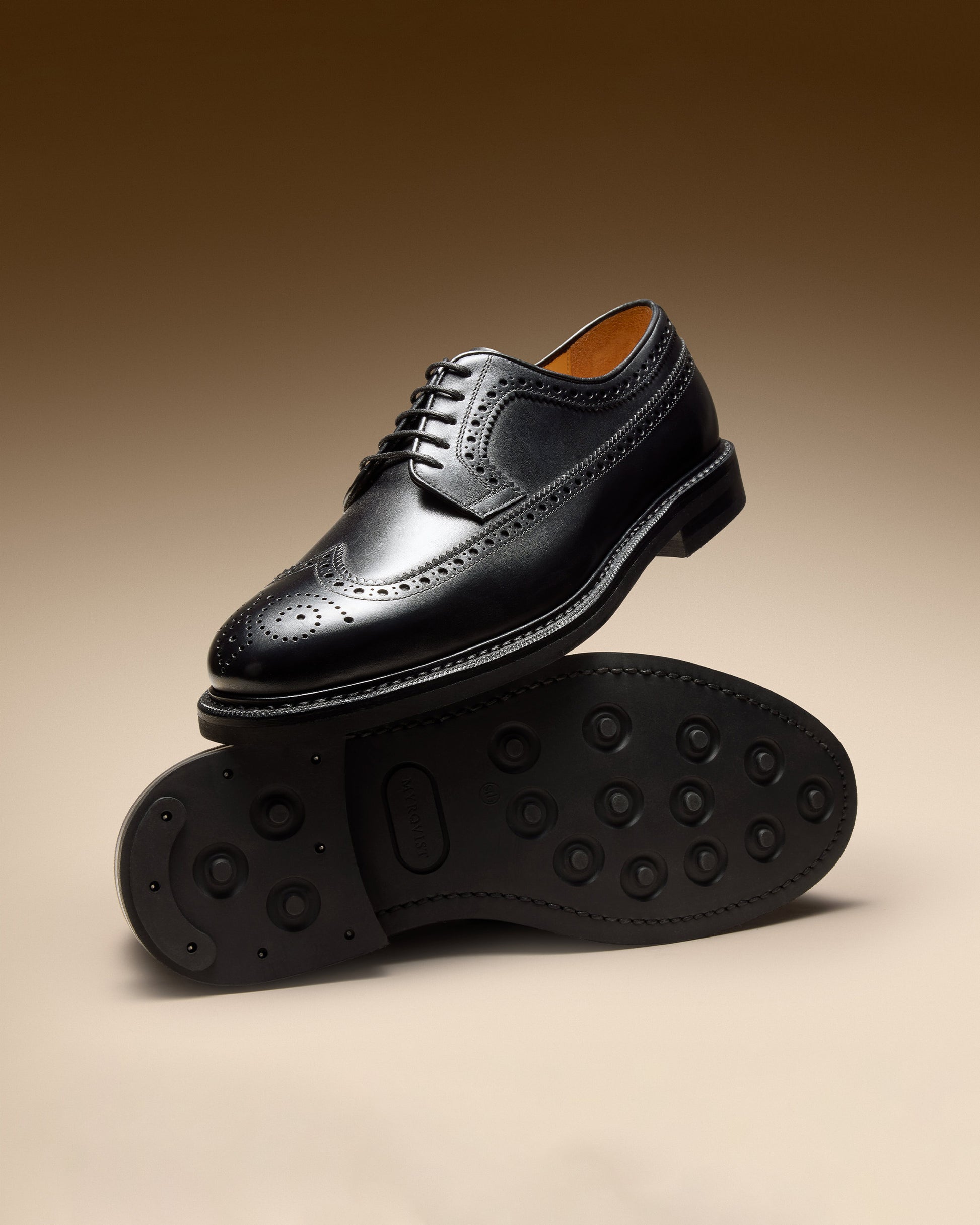 Side profile of Orust II – Black Calf brogue derby resting on its mate's studded rubber sole against a brown background.
