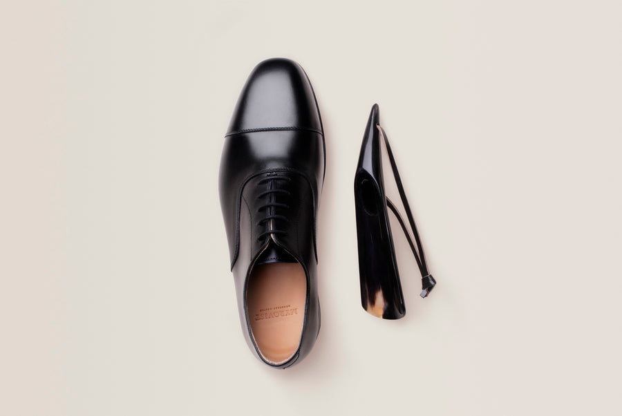 Top-down view of a laced black leather oxford beside a black leather belt on a pale background.