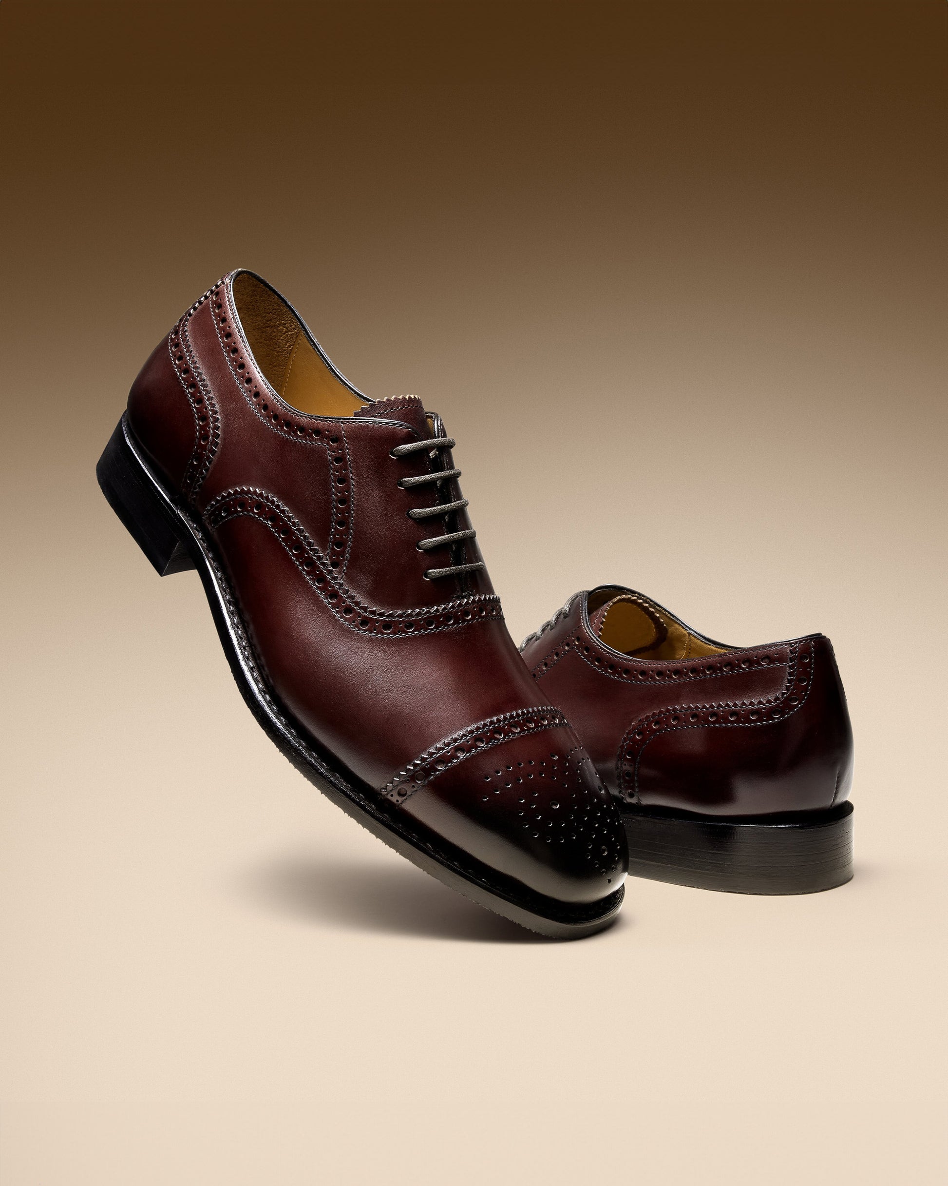 Studio shot of a pair of Strömsö II – Dark Mahogany Calf oxfords with brogue detailing, against a graduated tan background.