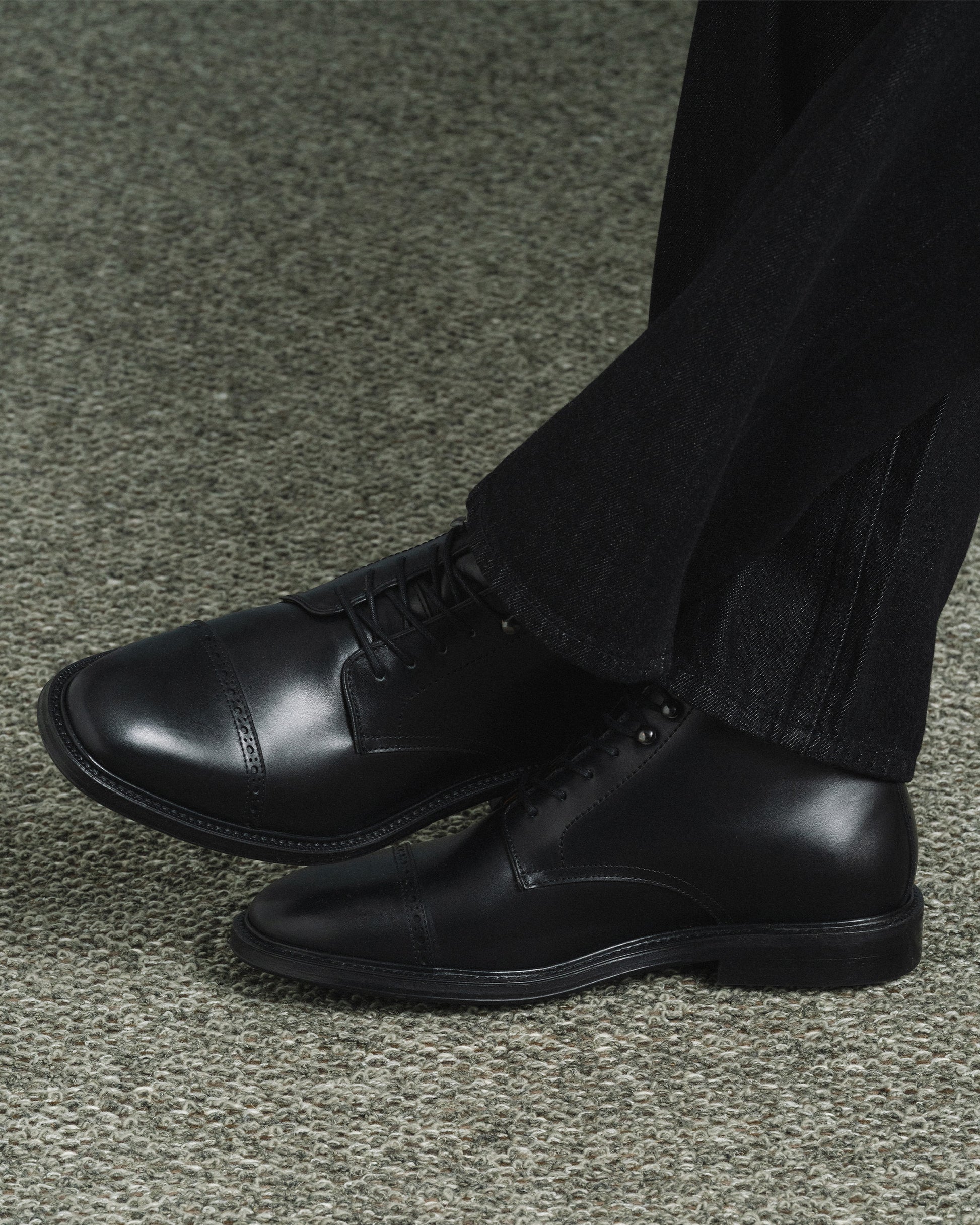 Tjärnö II in black calf worn with dark denim, highlighting the sleek cap toe, subtle broguing, and versatile design suited for both formal and casual settings.