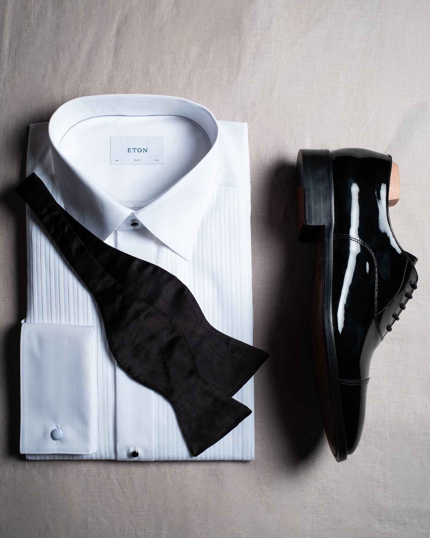 Une chemise de smoking blanche pliée avec un nœud papillon noir à gauche et, à droite, un seul richelieu en cuir verni noir présenté de profil, sur du lin naturel.