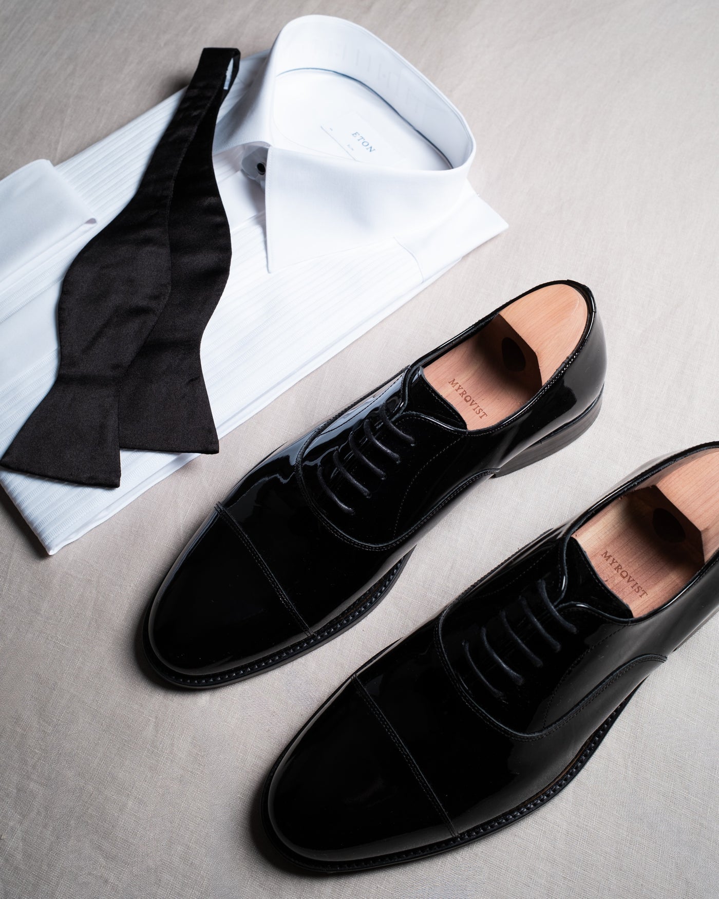 Une chemise de smoking blanche avec un nœud papillon noir dénoué à côté d'une paire de richelieus en cuir verni noir, photographiée d'en haut sur du lin naturel.
